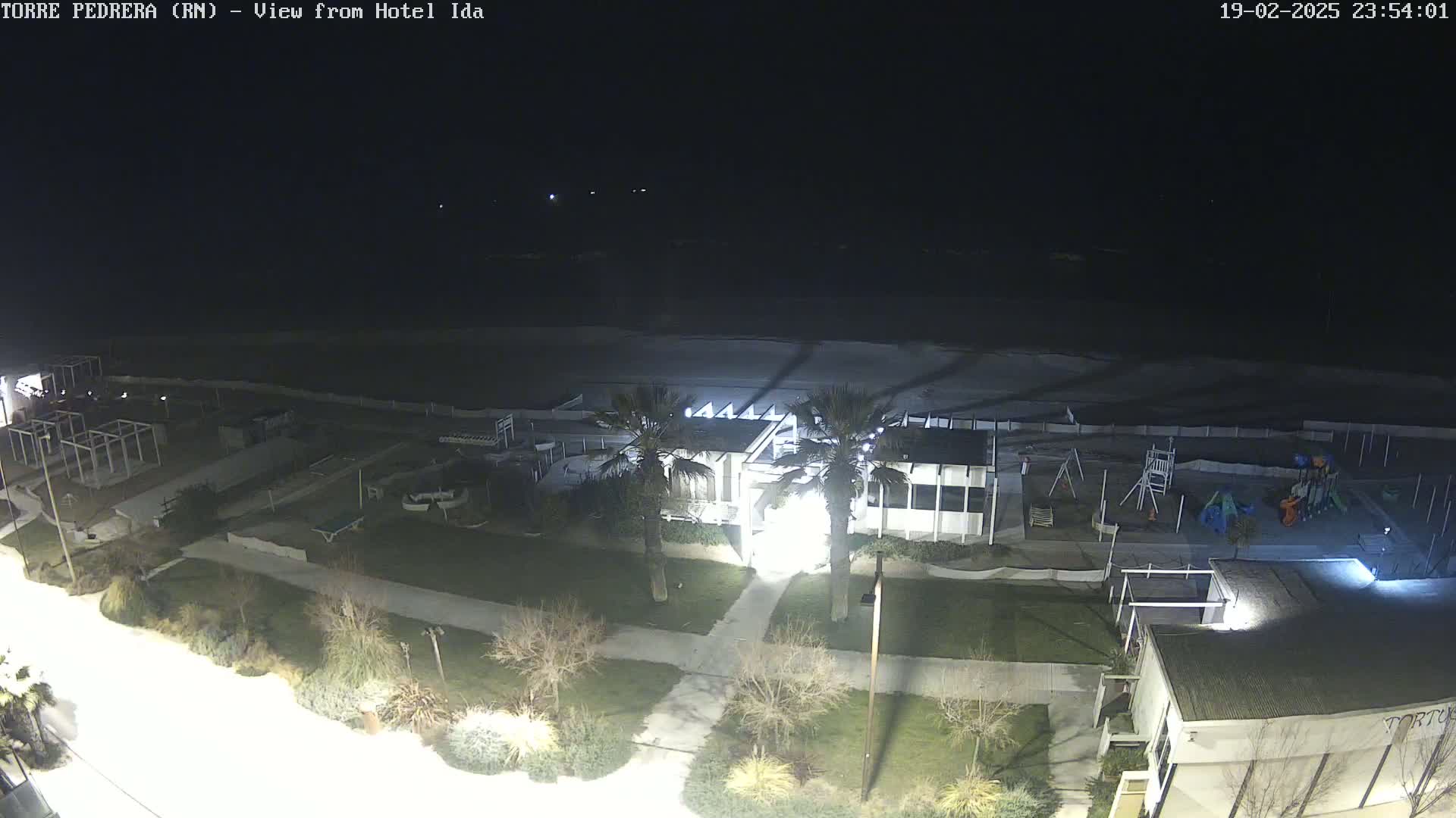 A nighttime aerial view shows a beach, a building with lights on, a playground, and sparse vegetation under a dark sky.