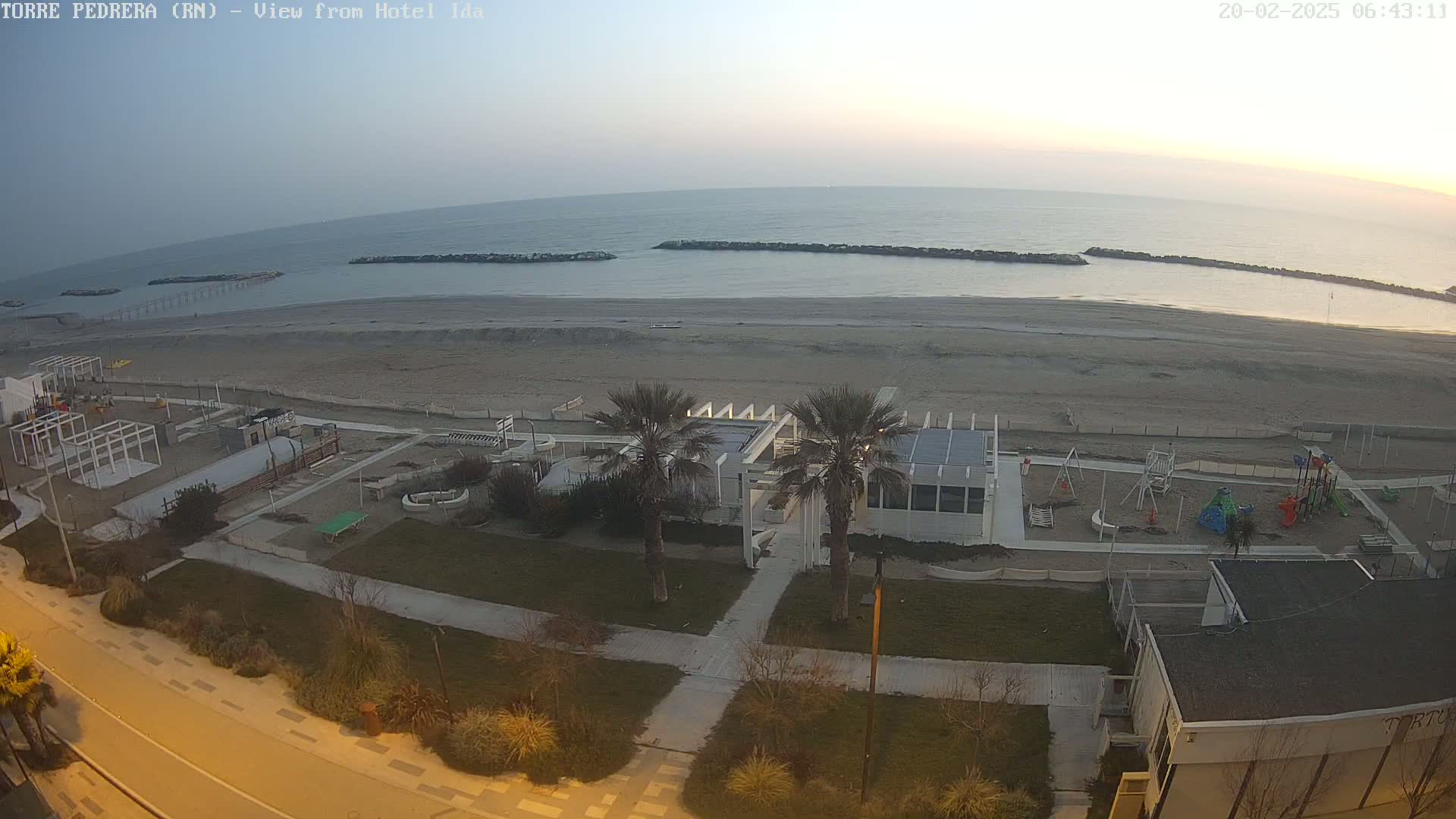 An aerial view shows a beach with calm, pale water under a clear, dawn sky, and a playground and beachfront buildings situated on the sand.