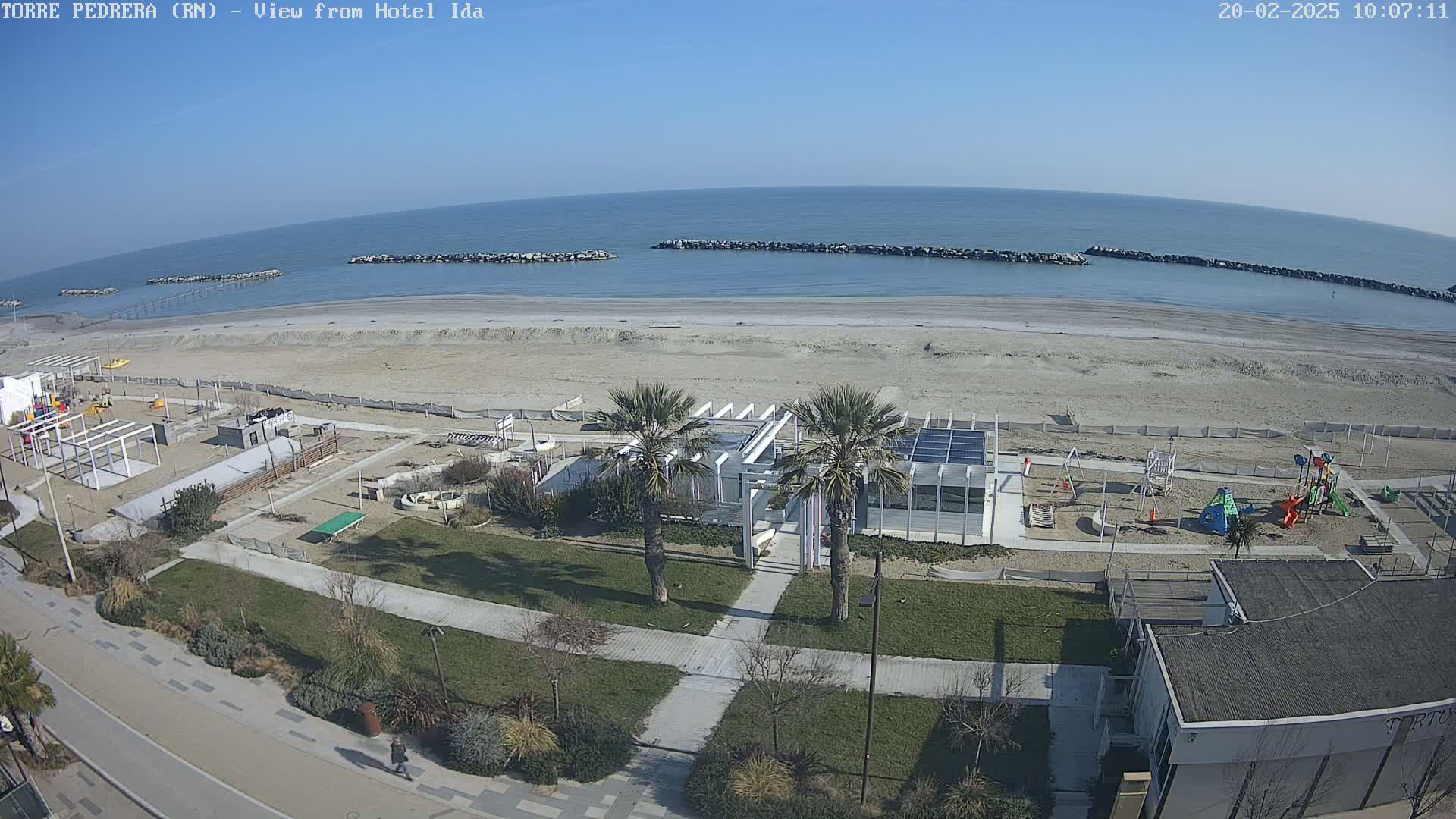 A sandy beach with calm, blue water under a clear, sunny sky is bordered by a paved walkway, grassy areas with palm trees, and children’s playgrounds, all viewed from above.