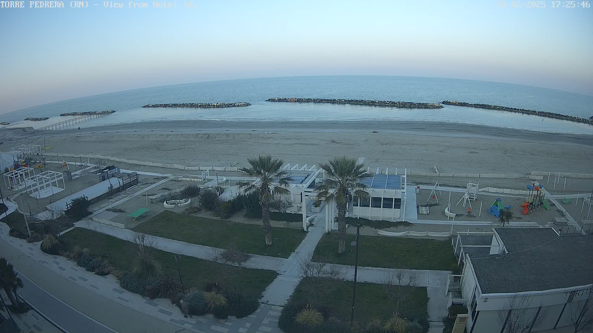 An aerial view of a beach with calm water under a twilight sky shows a sandy shore with a playground, palm trees, and a beachfront building.