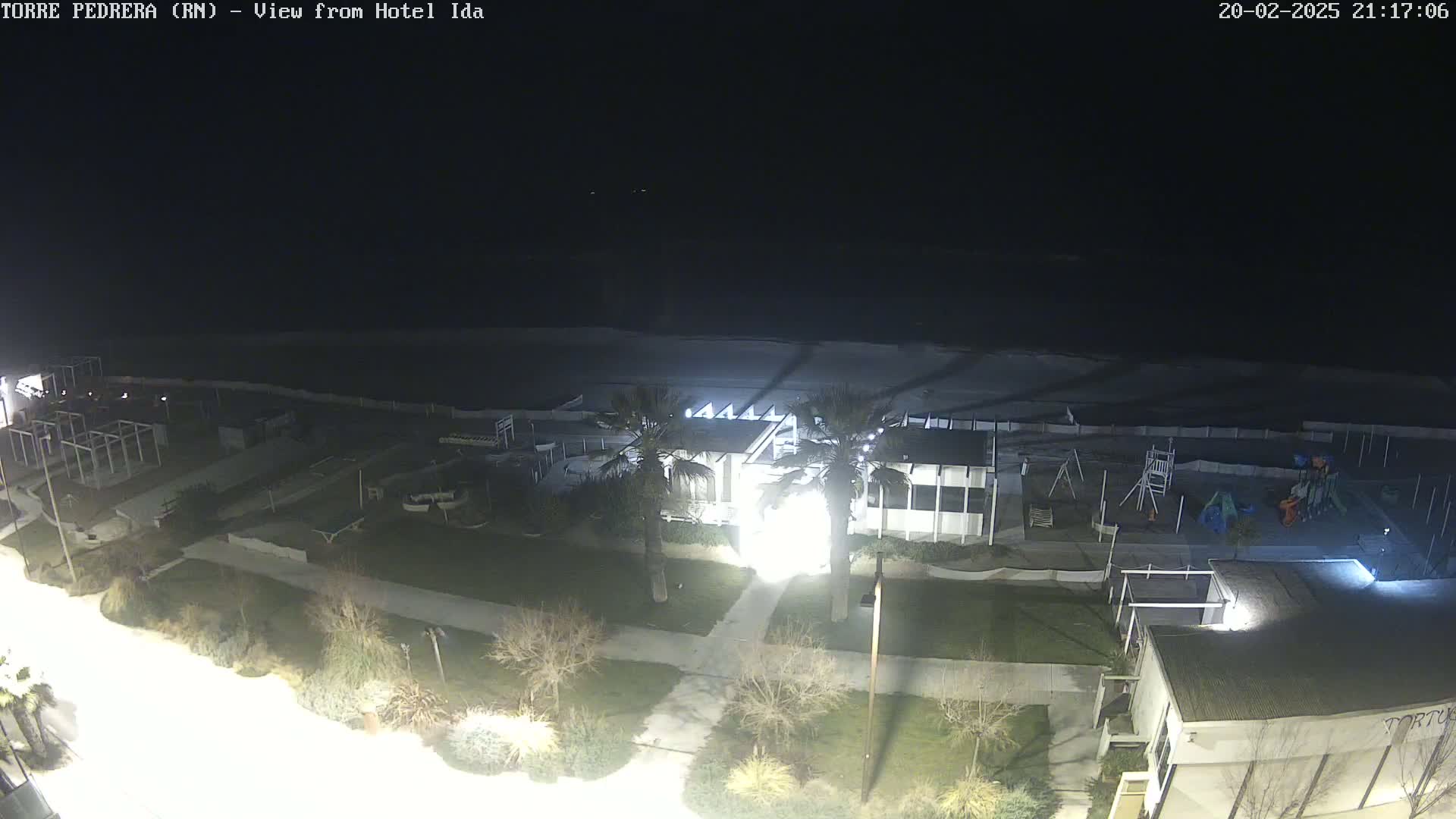 A nighttime view of a beachside area shows a building with palm trees and playground equipment, a dark ocean, and a mostly empty beach.