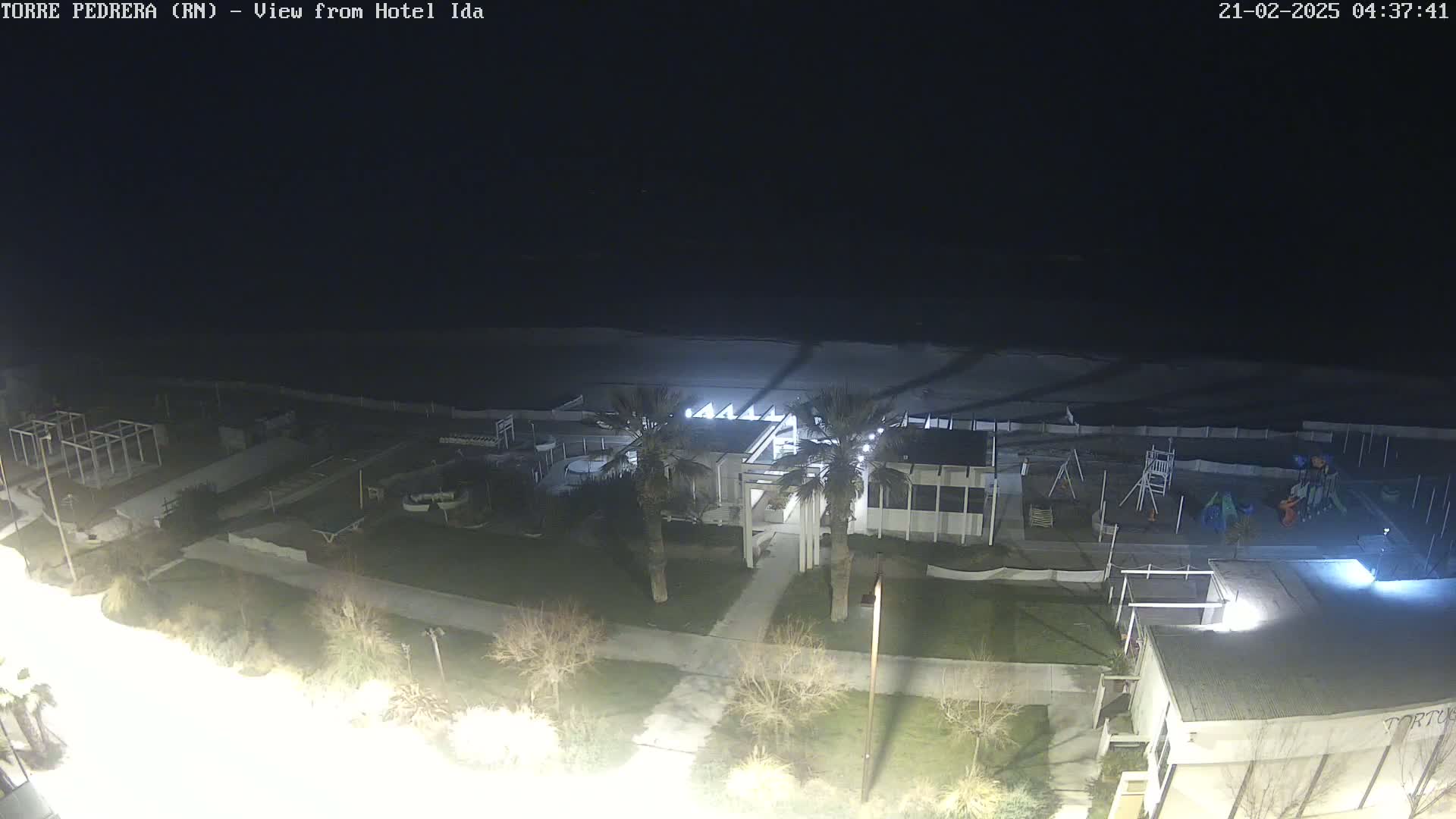 A nighttime, overhead view of a beach area shows a building with palm trees in front of it, a playground, and a dark ocean under a clear, dark sky.