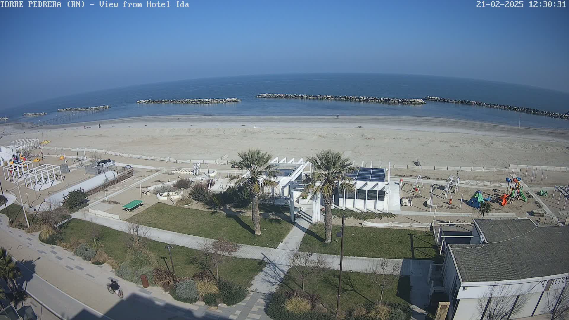 A sunny day overlooking a sandy beach with calm water, breakwaters, a playground, and a beachfront building with palm trees.