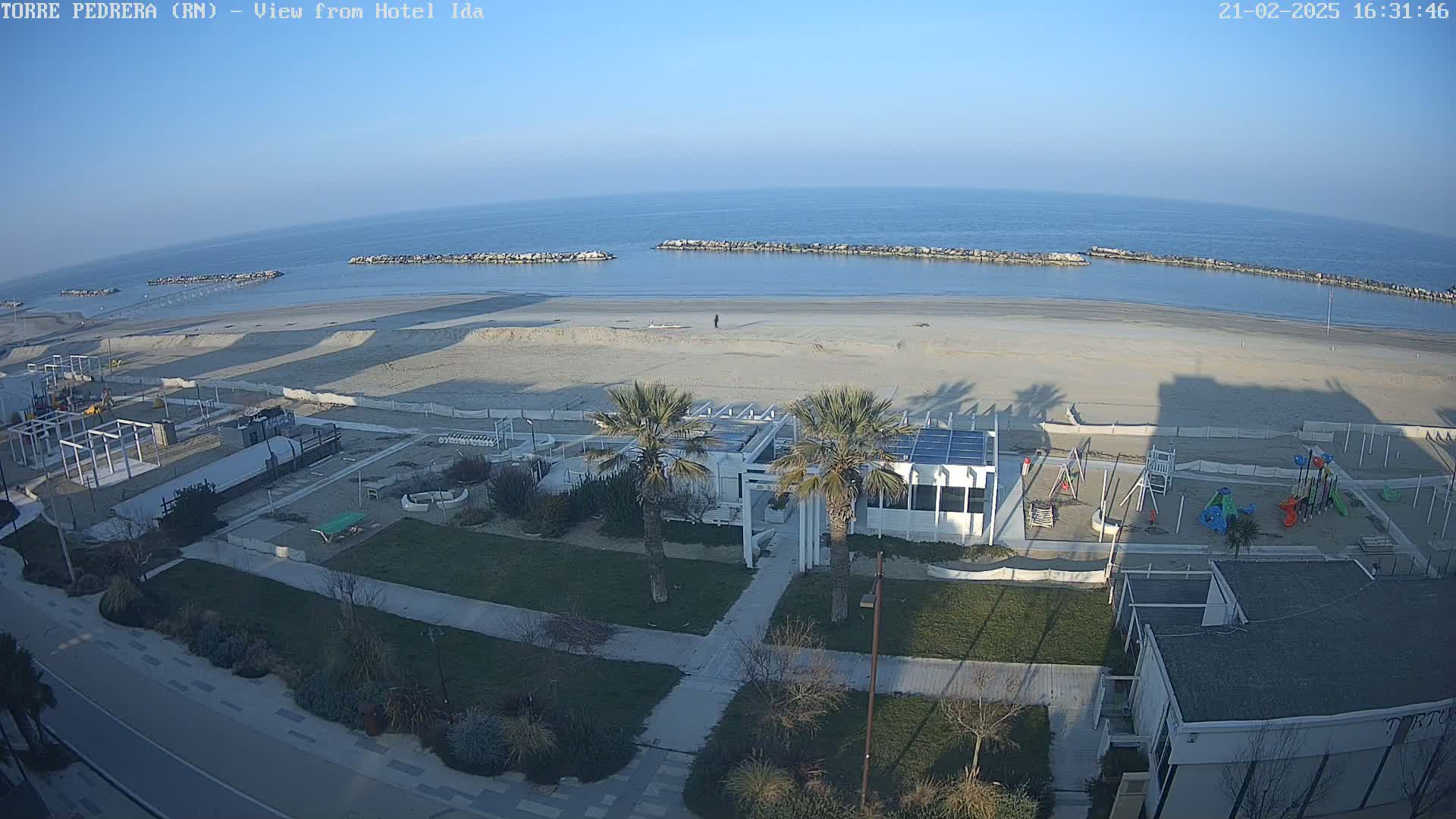 An aerial view shows a beach with calm, clear water under a clear blue sky, adjacent to a beachfront area with palm trees, buildings, and a playground.