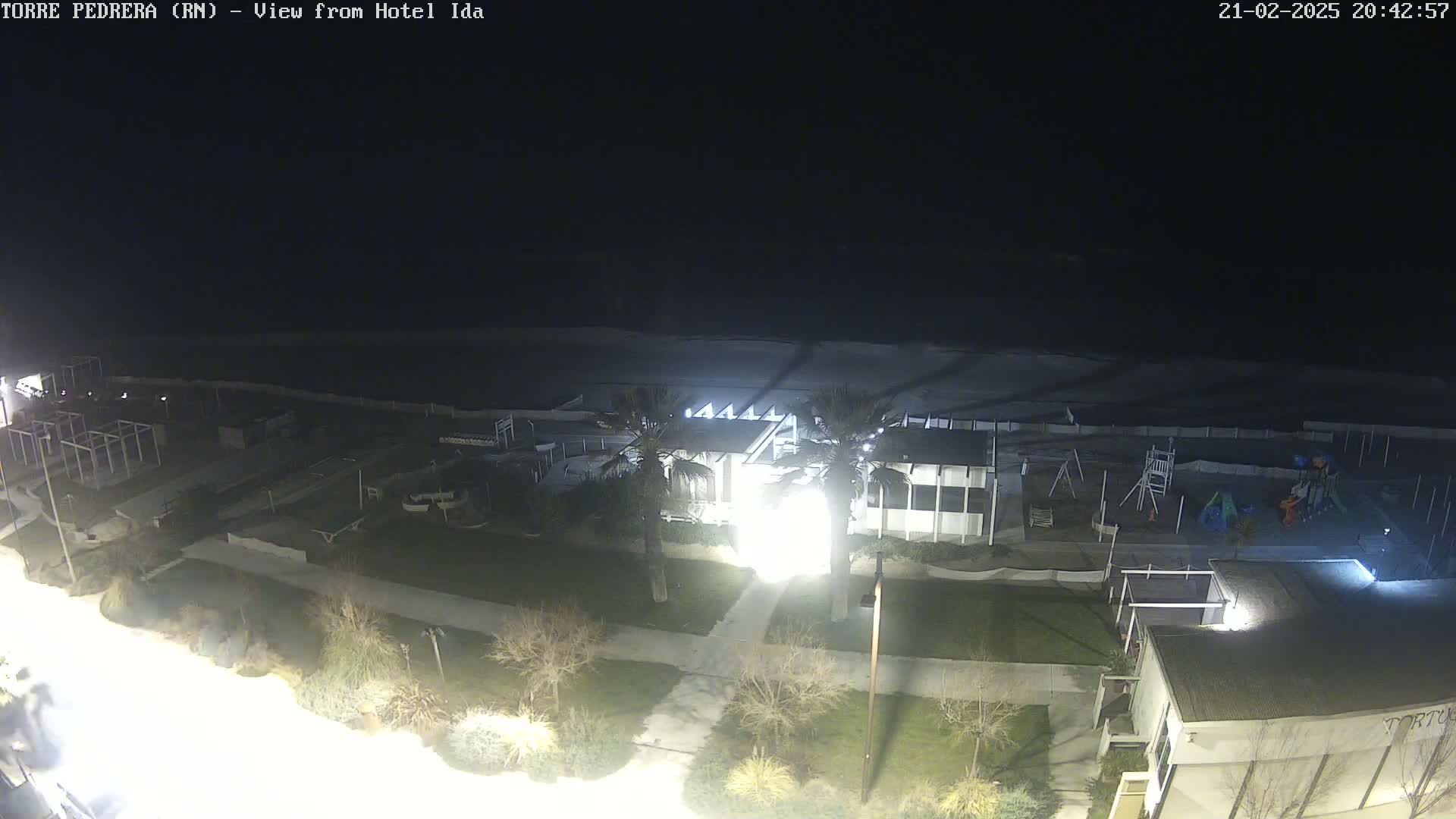 A nighttime view of a beachside area shows a building with lights on, a playground, and a dark ocean under a clear sky.