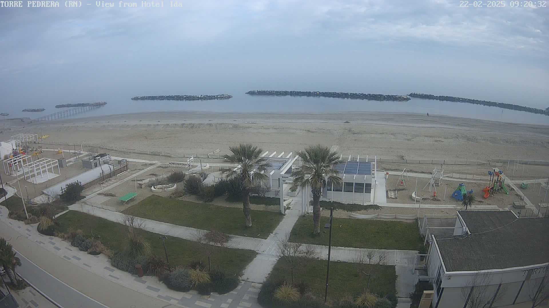 An overcast day reveals a beach with calm water, a playground, and a beachfront building with palm trees, all viewed from above.