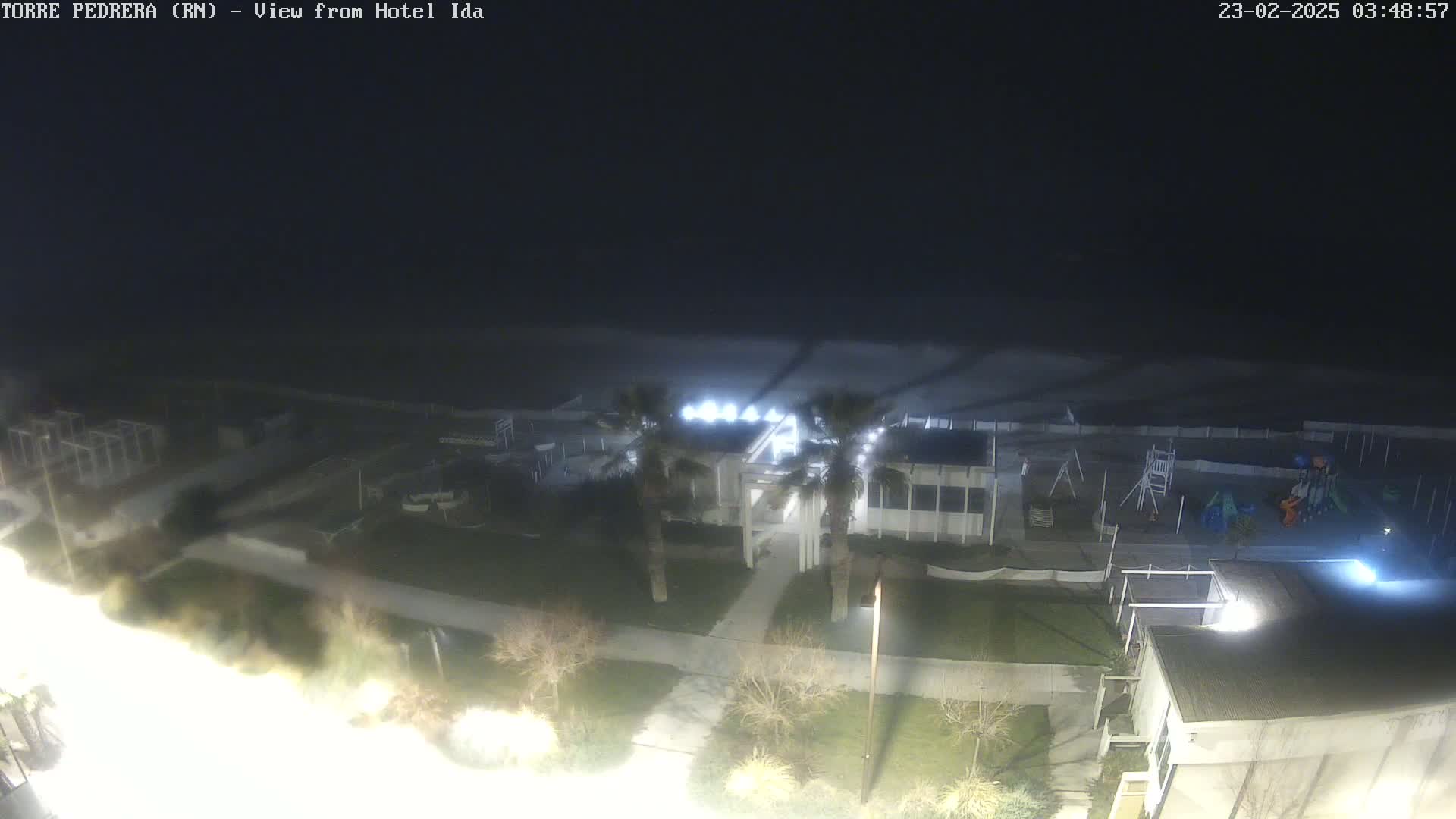A nighttime aerial view of a beach with a building, palm trees, and a playground, under dark, cloudy skies.