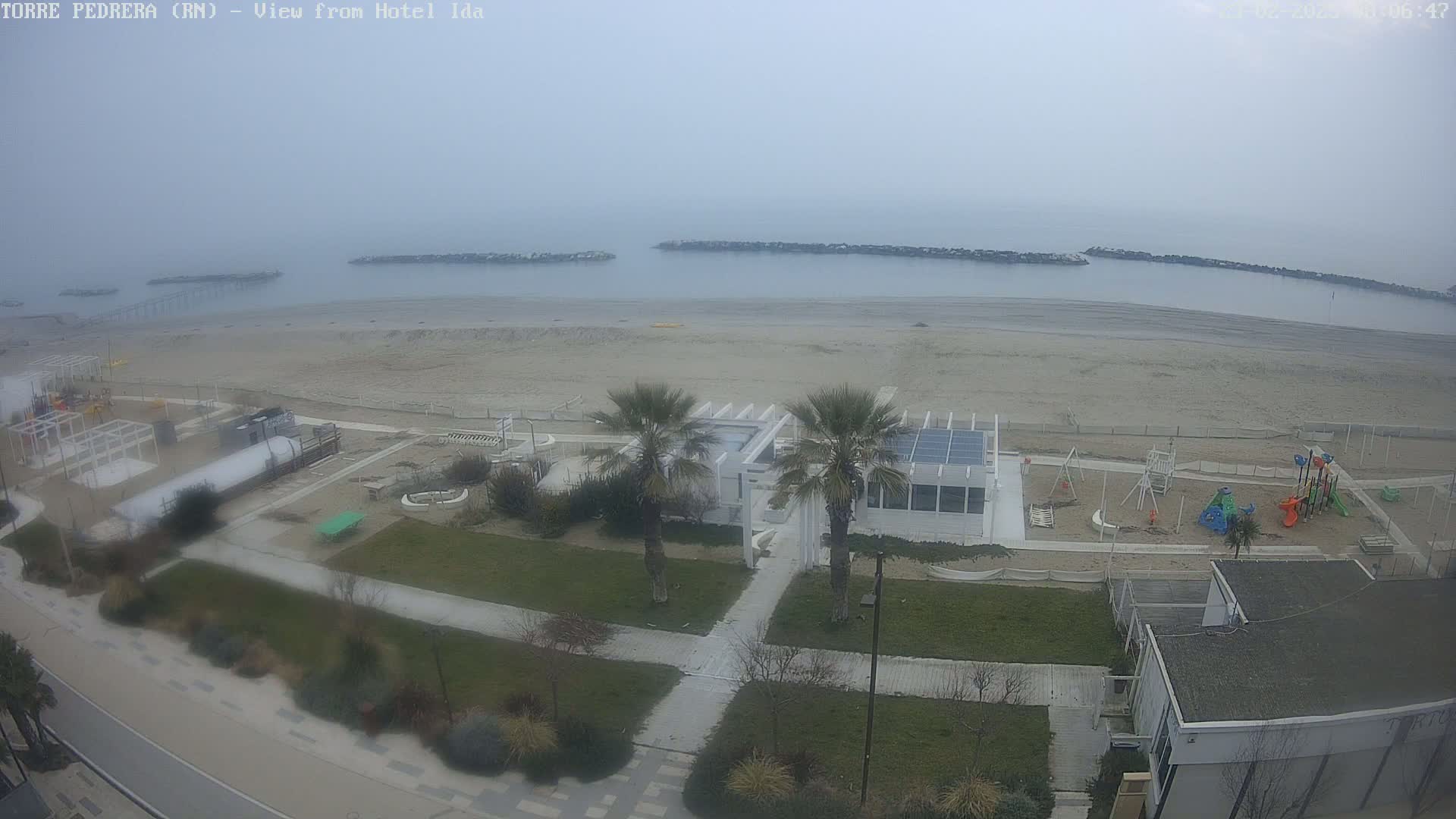 A hazy coastal view shows a sandy beach, a playground, palm trees, and buildings near the ocean on an overcast day.