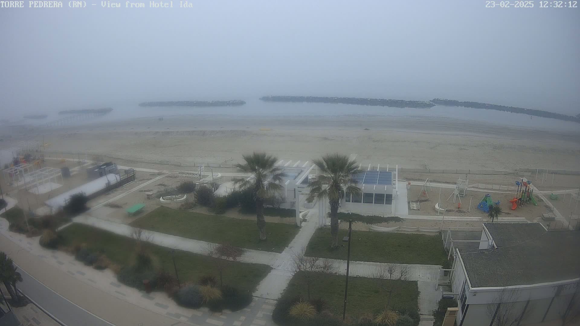 A foggy seaside view shows a sandy beach, a playground, palm trees, and buildings near a calm body of water.