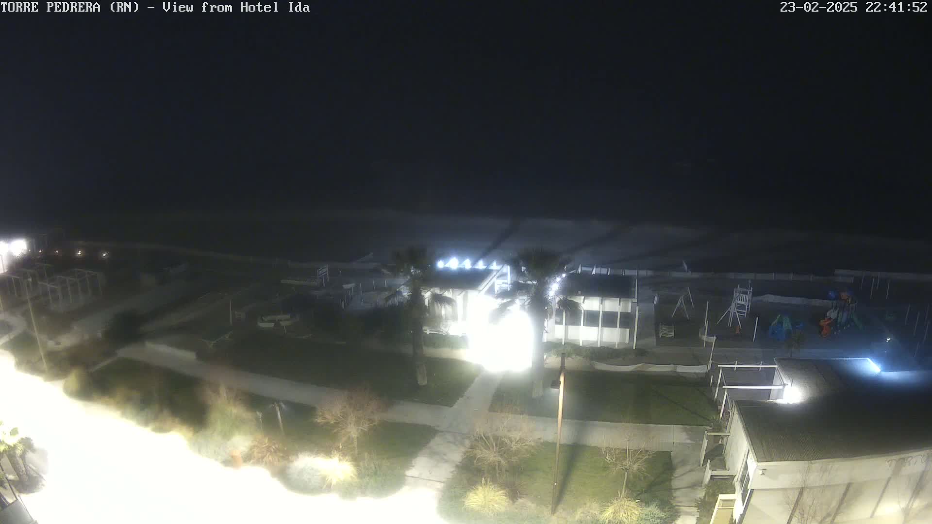 A nighttime view of a beachside area shows a building, playground, and palm trees illuminated under dark skies.