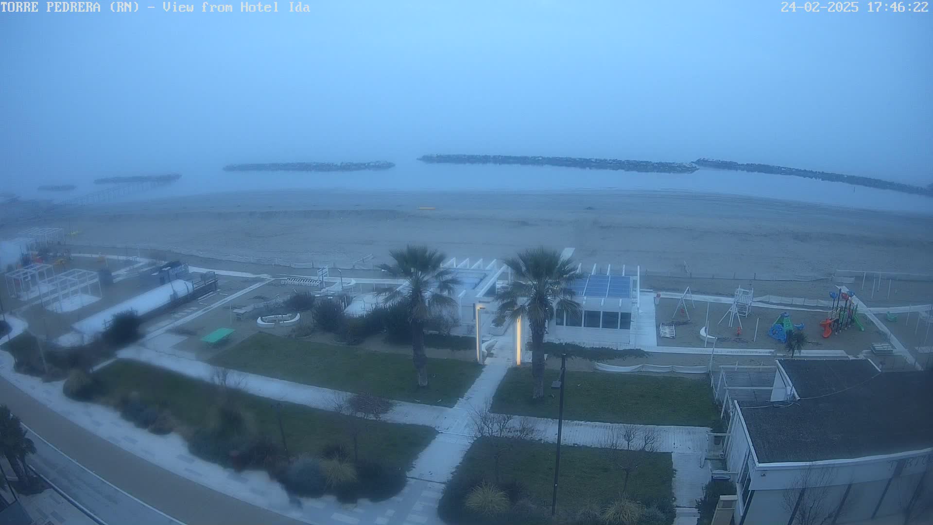 A foggy coastal scene shows a beach, a playground, beachfront buildings, and palm trees.