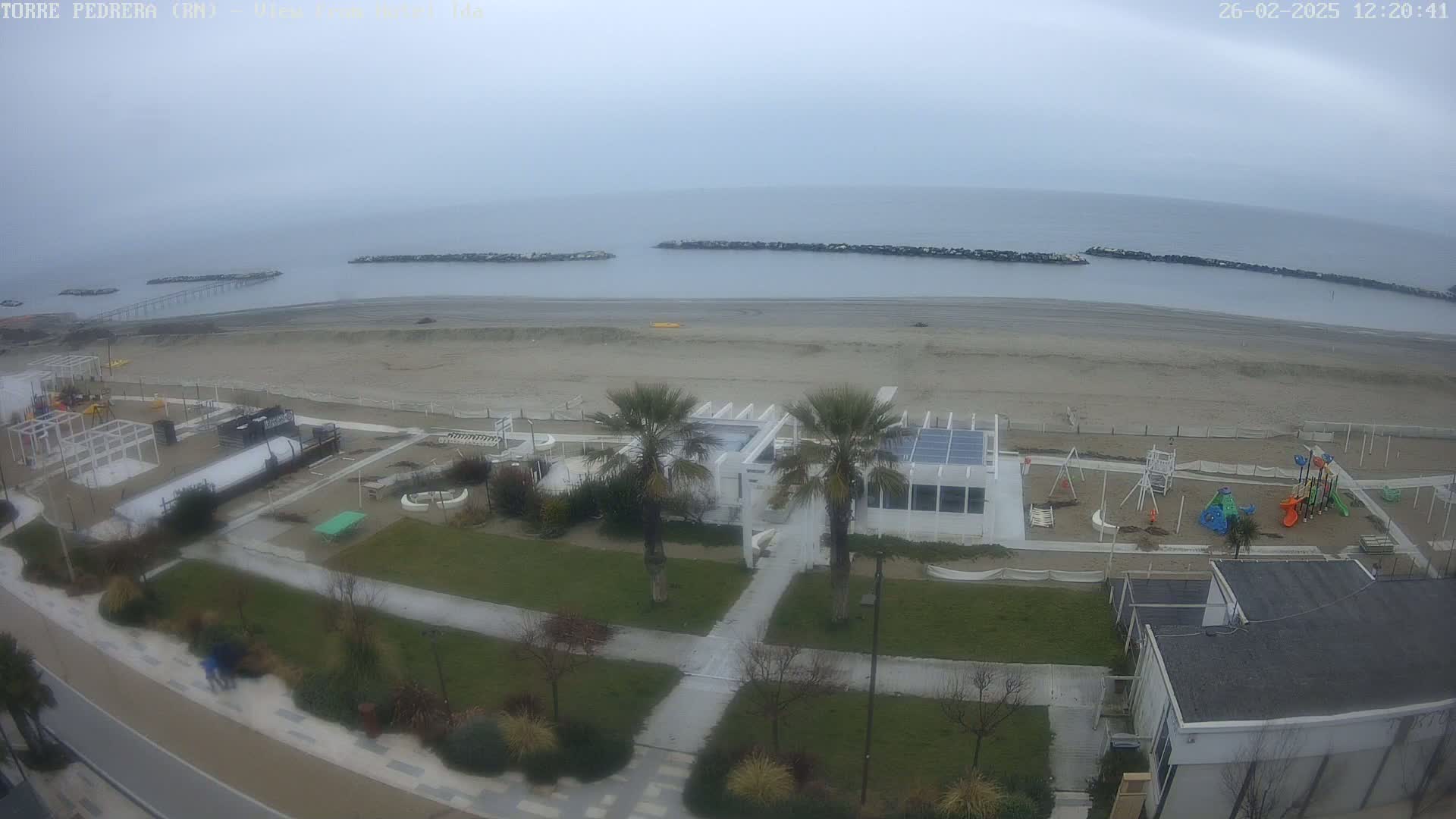An overcast day shows a sandy beach with a playground, beachfront buildings, and palm trees, viewed from above.
