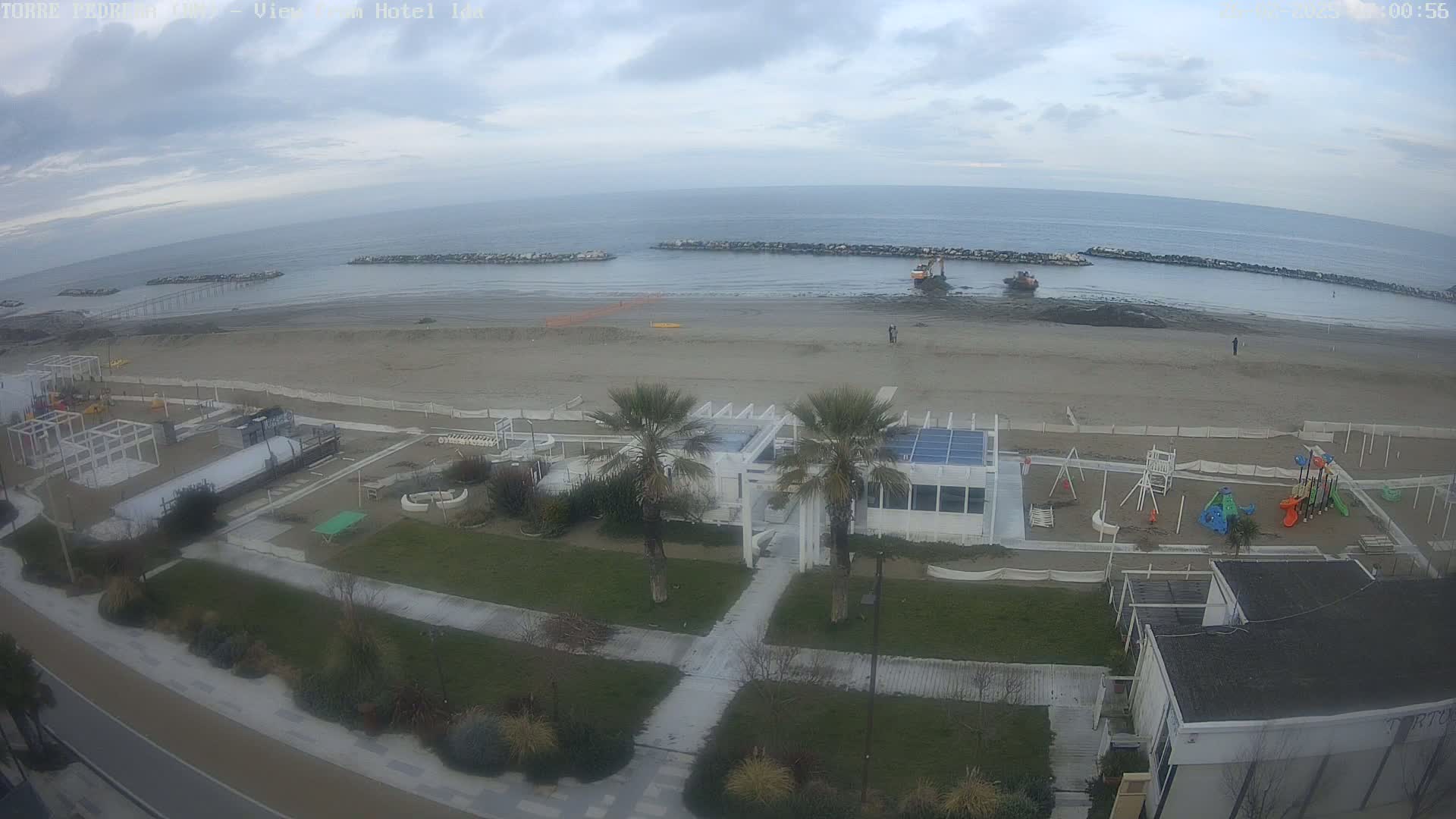 A beach scene on an overcast day shows a sandy beach with calm water, beachside structures, playground equipment, and some construction equipment on the water's edge.