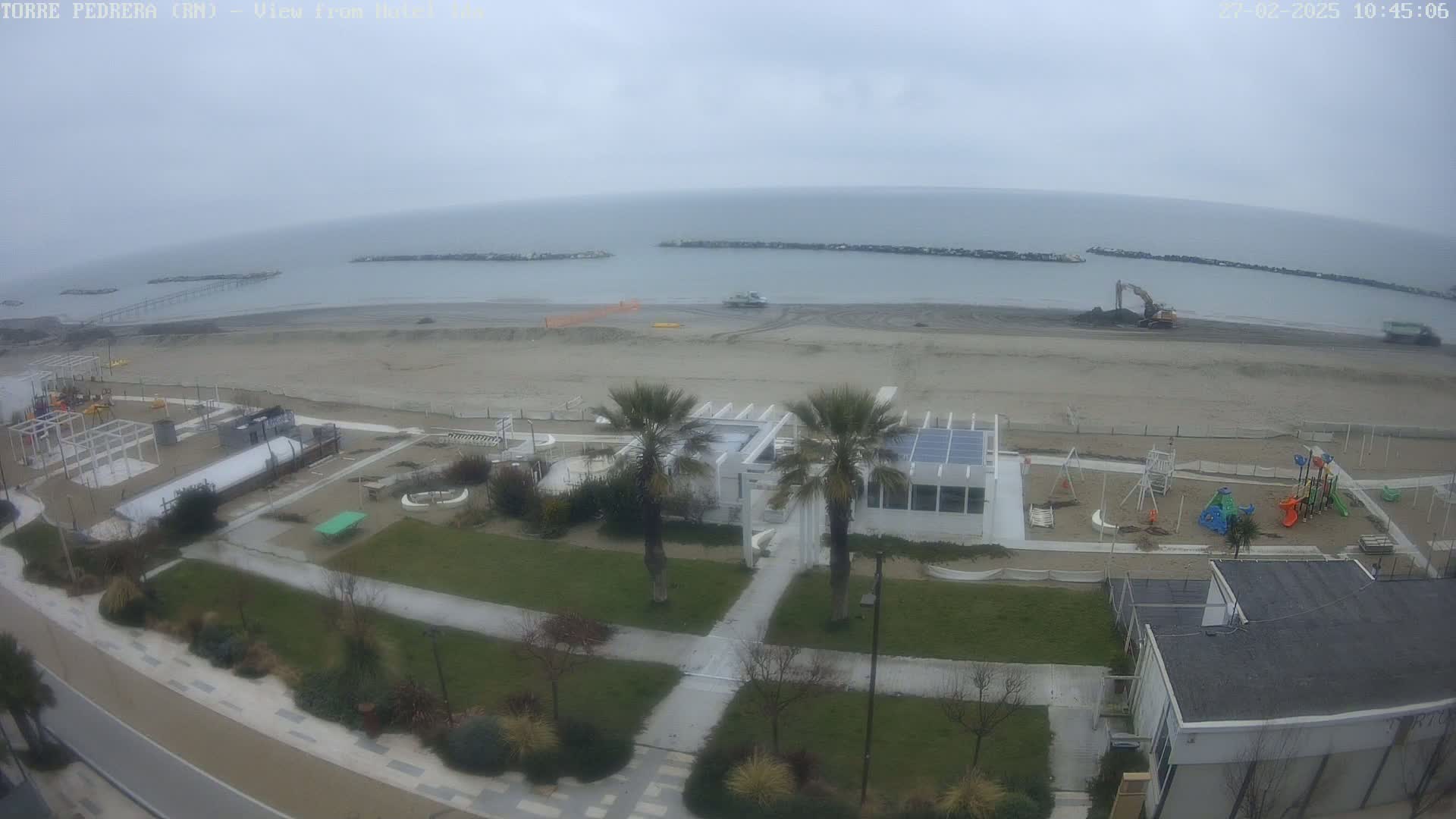 An overcast day shows an aerial view of a beach with construction equipment, a playground, palm trees, and buildings.
