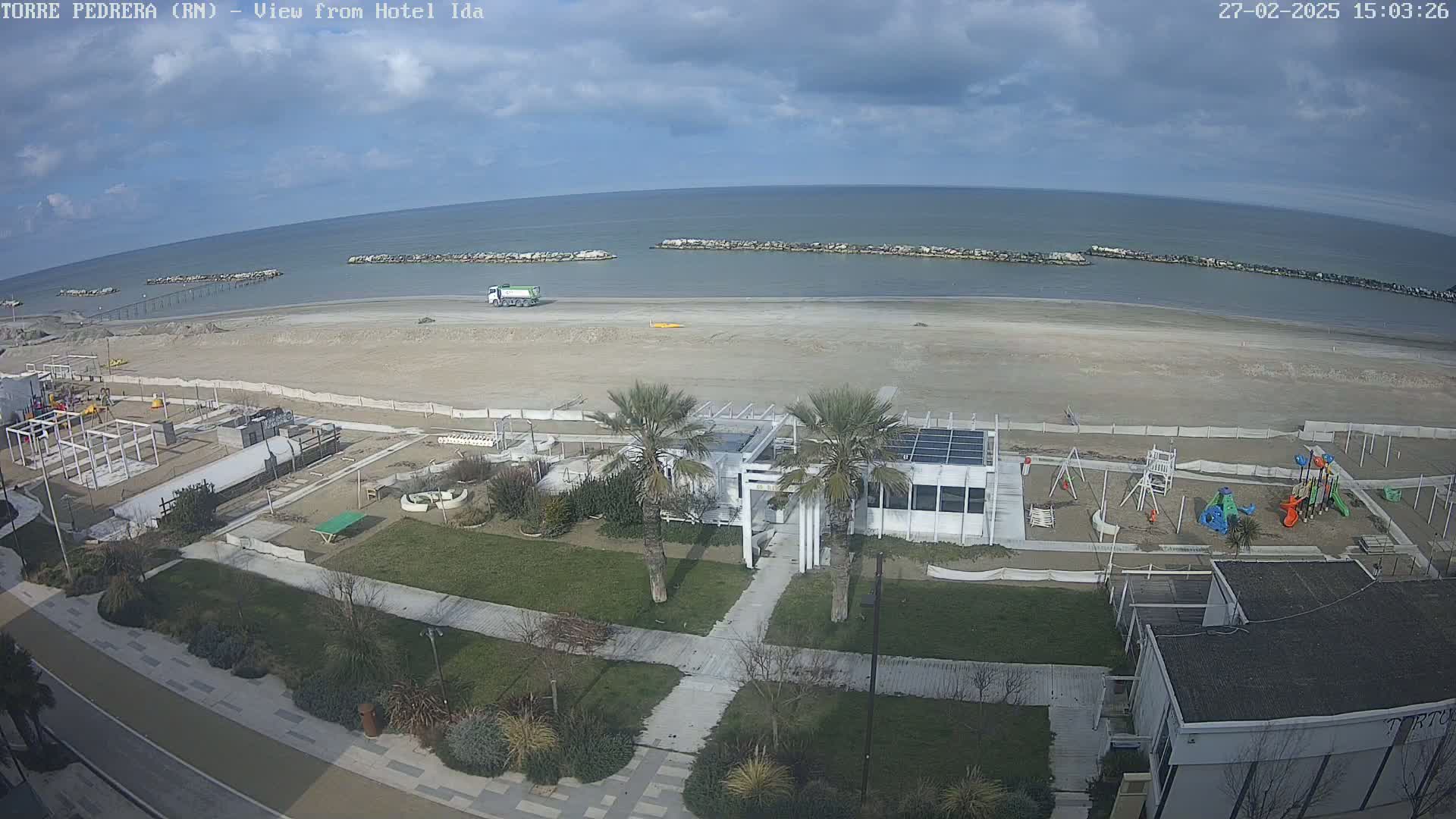 An aerial view shows a mostly empty sandy beach under a partly cloudy sky, with a playground, some buildings, and palm trees along the beachfront.