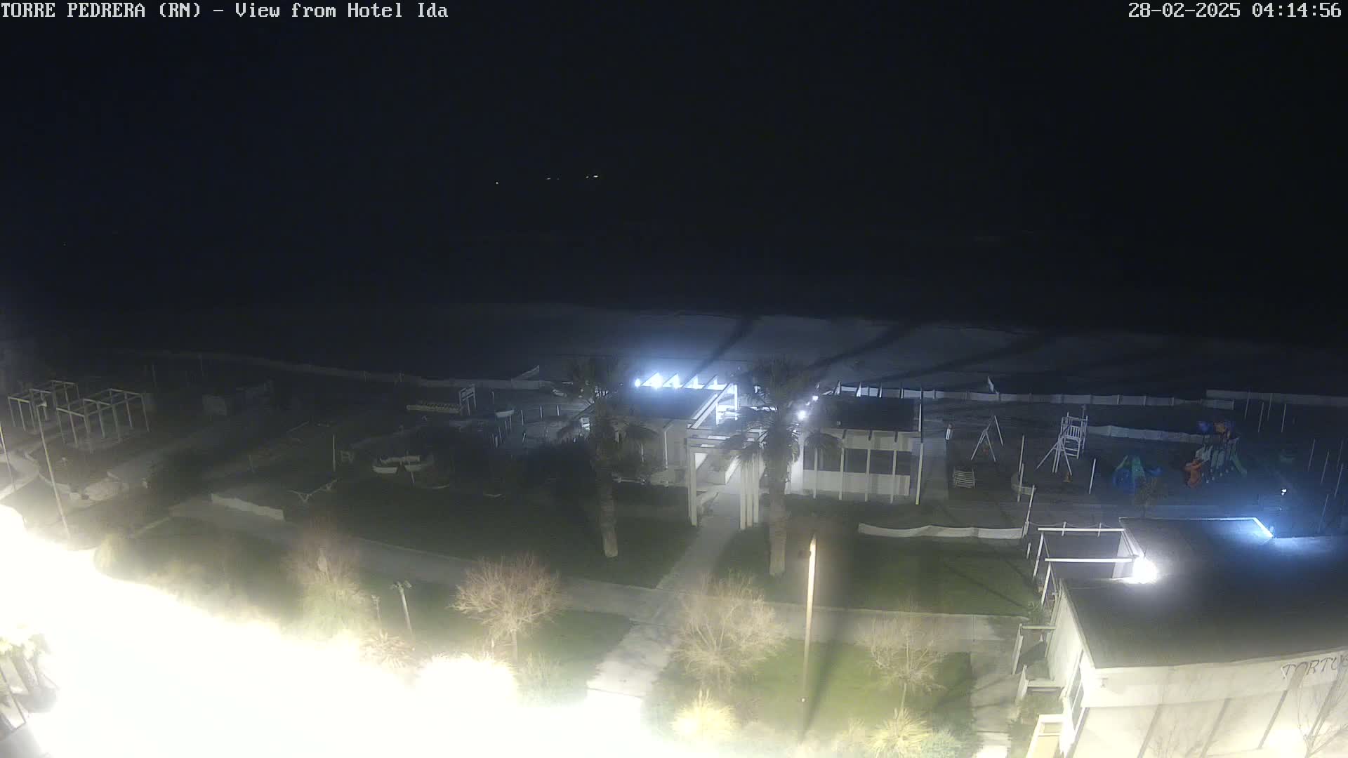 A nighttime aerial view shows a beach, a playground, a building with lights on, and surrounding landscaping.