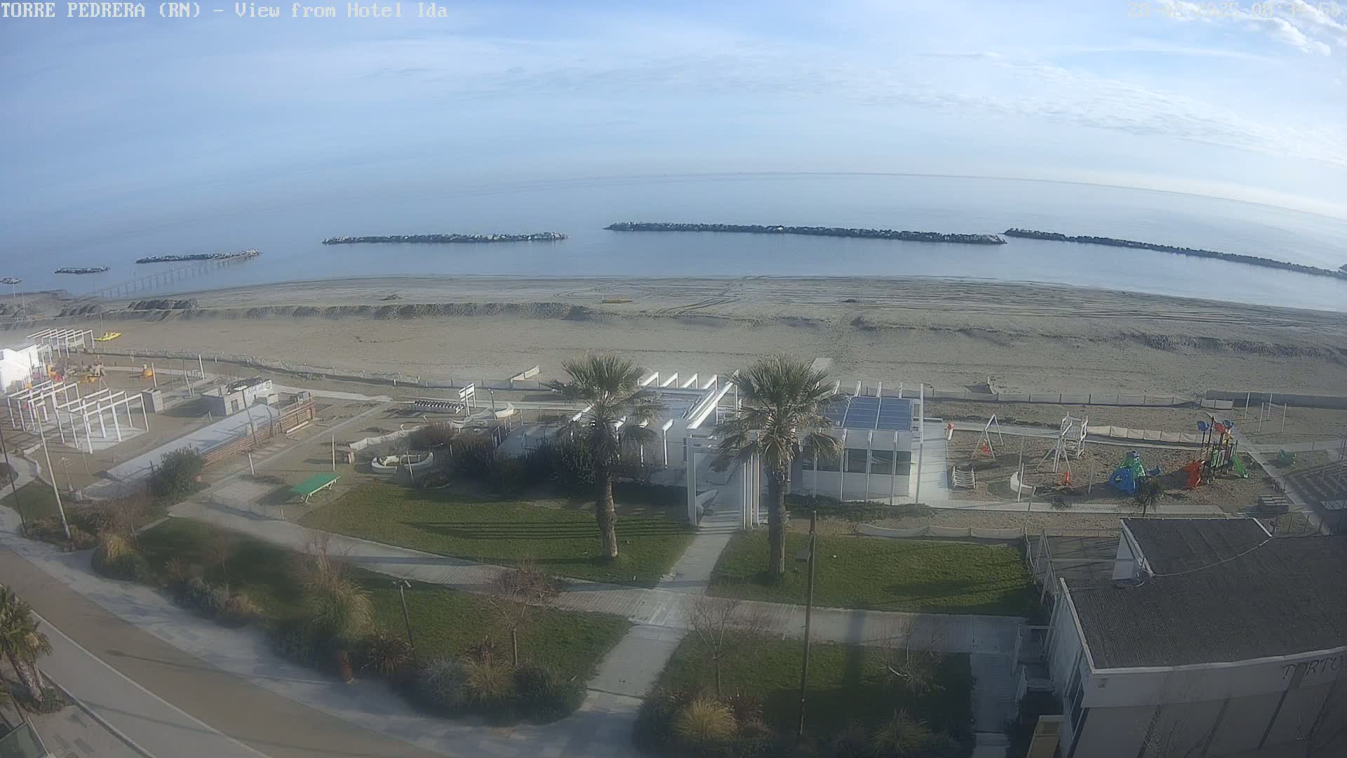 A sandy beach stretches along a calm, clear ocean under a mostly sunny sky, with a playground and a white building visible near the shore.