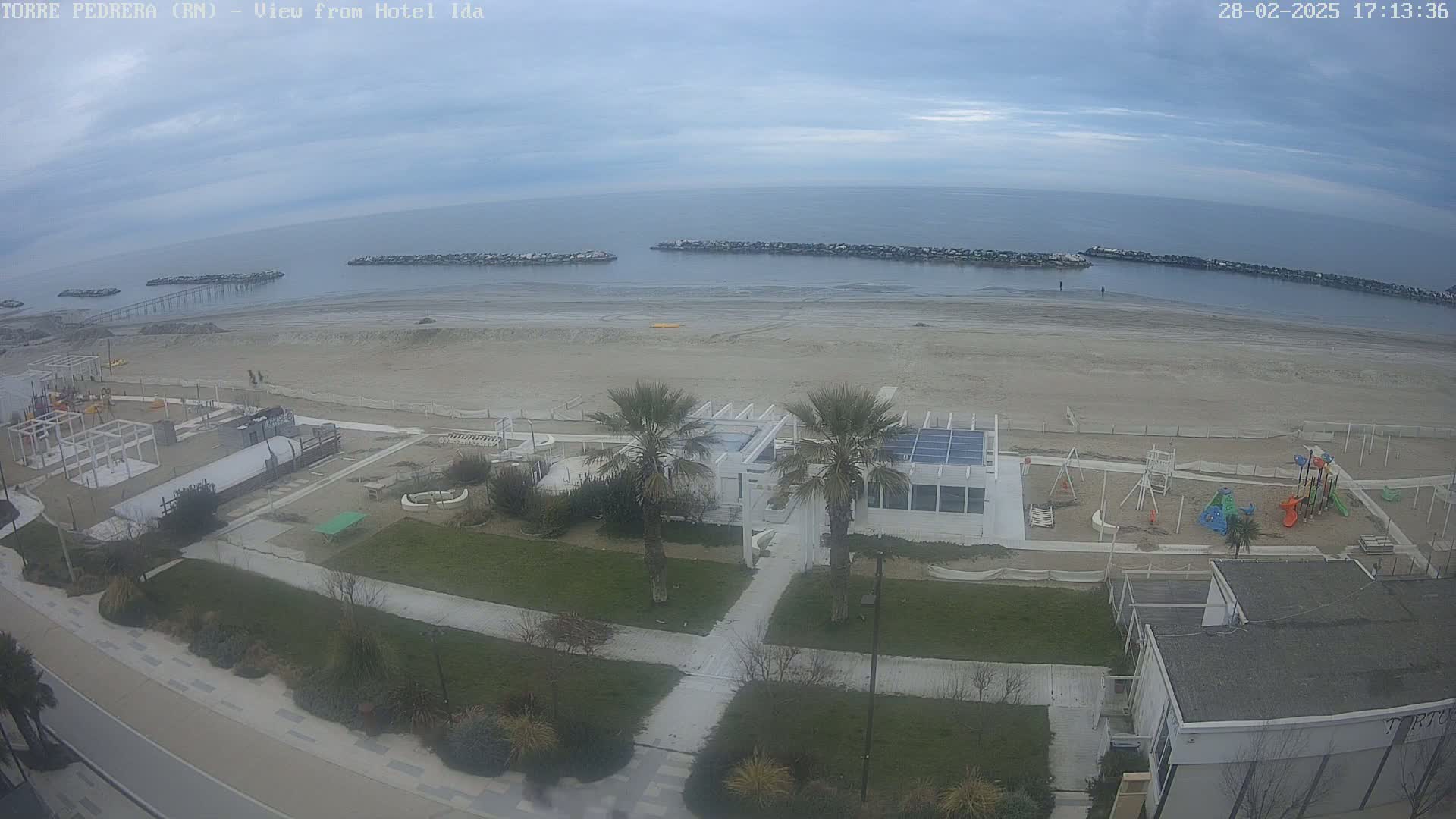 An overcast day reveals a sandy beach with a playground and beachside structures, all overlooked by palm trees and a building.