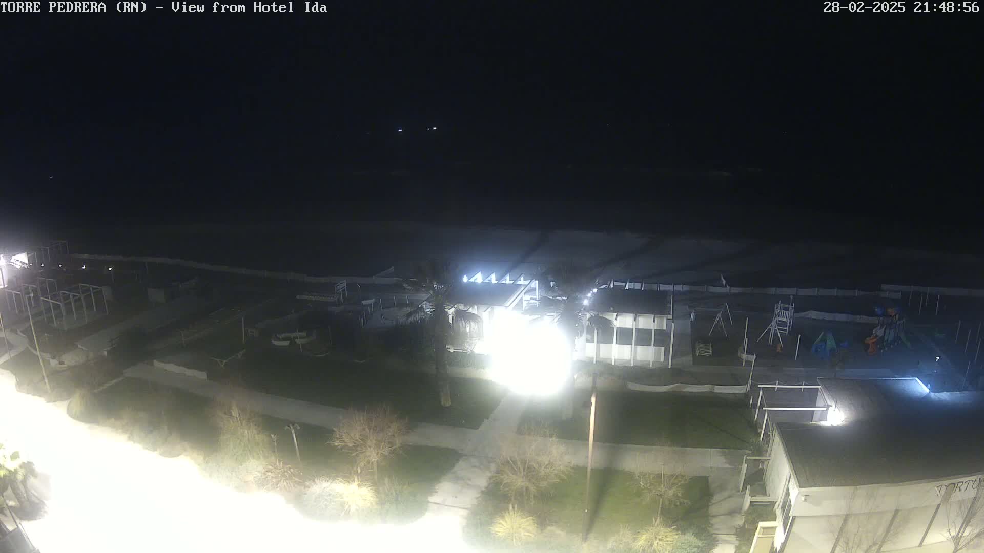 A nighttime, overhead view of a beach area with buildings, a playground, and palm trees, under a dark sky.