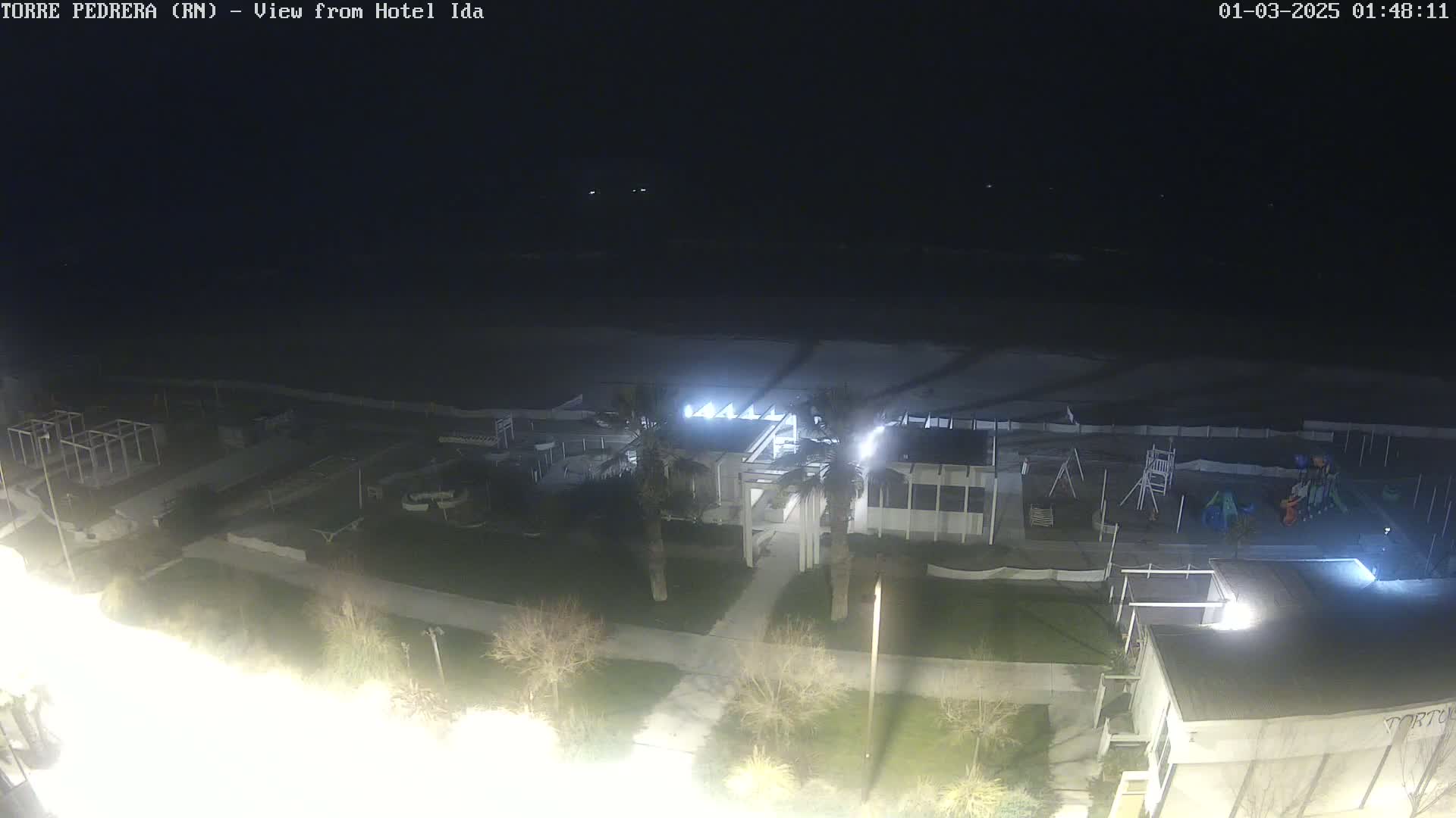 A nighttime, high-angle view shows a mostly empty beach, a playground, and a building complex illuminated by artificial lights.