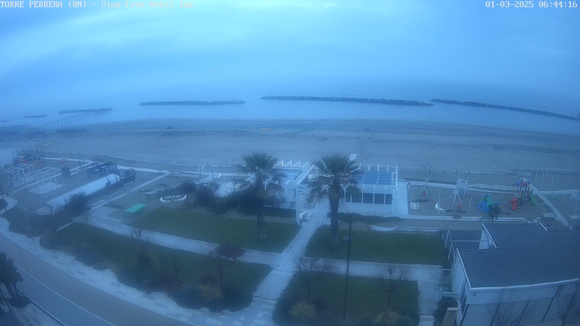 A high-angle, long shot shows a beach with calm water under an overcast sky, featuring a playground, palm trees, and buildings near the shoreline.