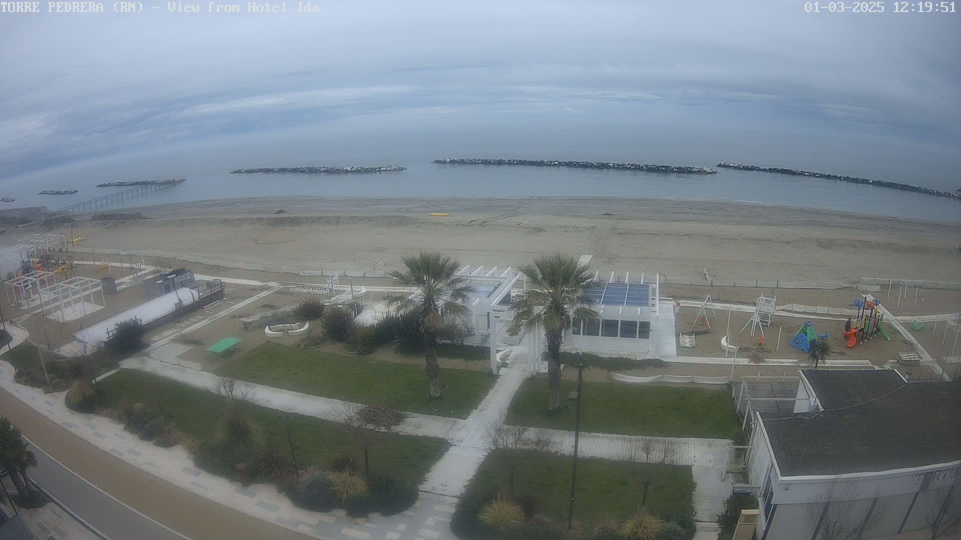 An aerial view shows a beach with calm, overcast skies, a playground, and palm trees near a building.