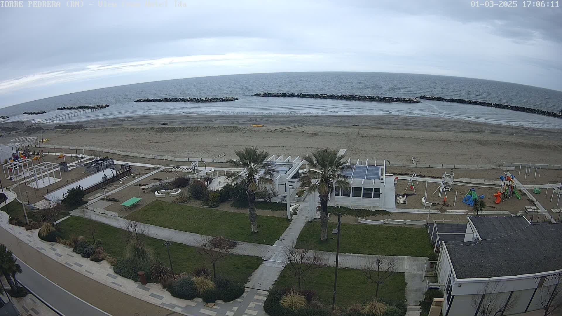 An overcast day shows a sandy beach with a calm sea,  adjacent to a grassy area with a playground, palm trees, and a white building, all viewed from an elevated perspective.