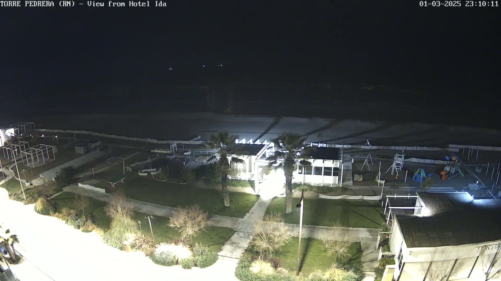 A nighttime view of a beach with a playground, some buildings, and palm trees, under a dark sky.