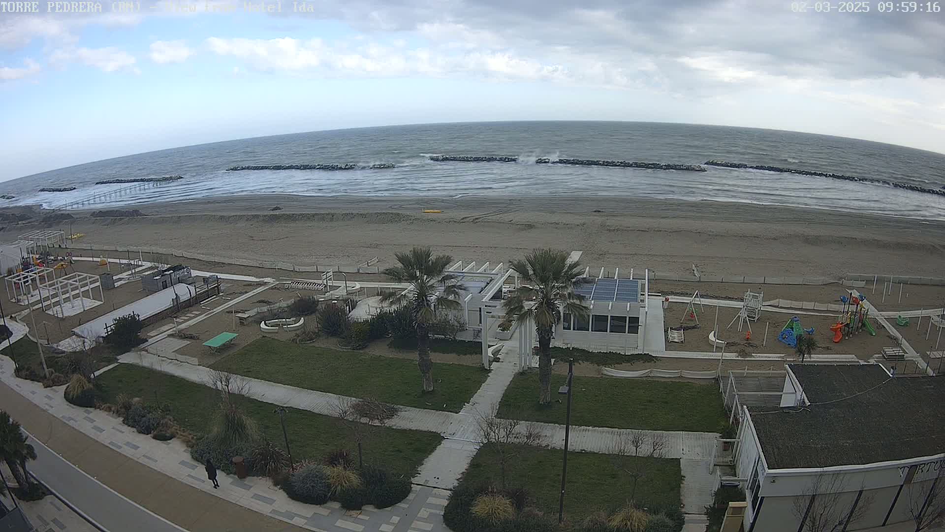 An overcast day shows a beach with a playground and beachfront building, and a choppy sea with waves breaking against rock barriers.