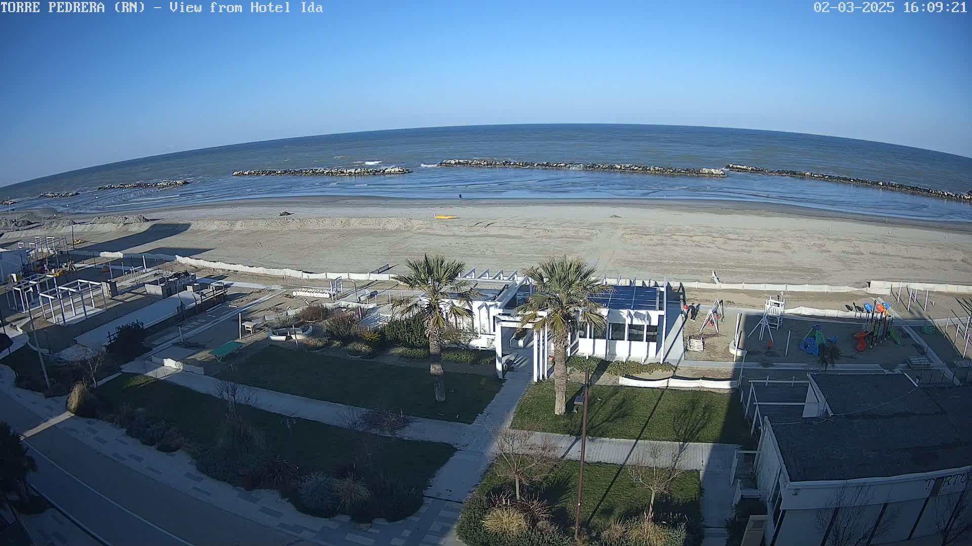 A sandy beach with calm ocean waves under a clear blue sky stretches behind a beachfront area with a playground, palm trees, and buildings.