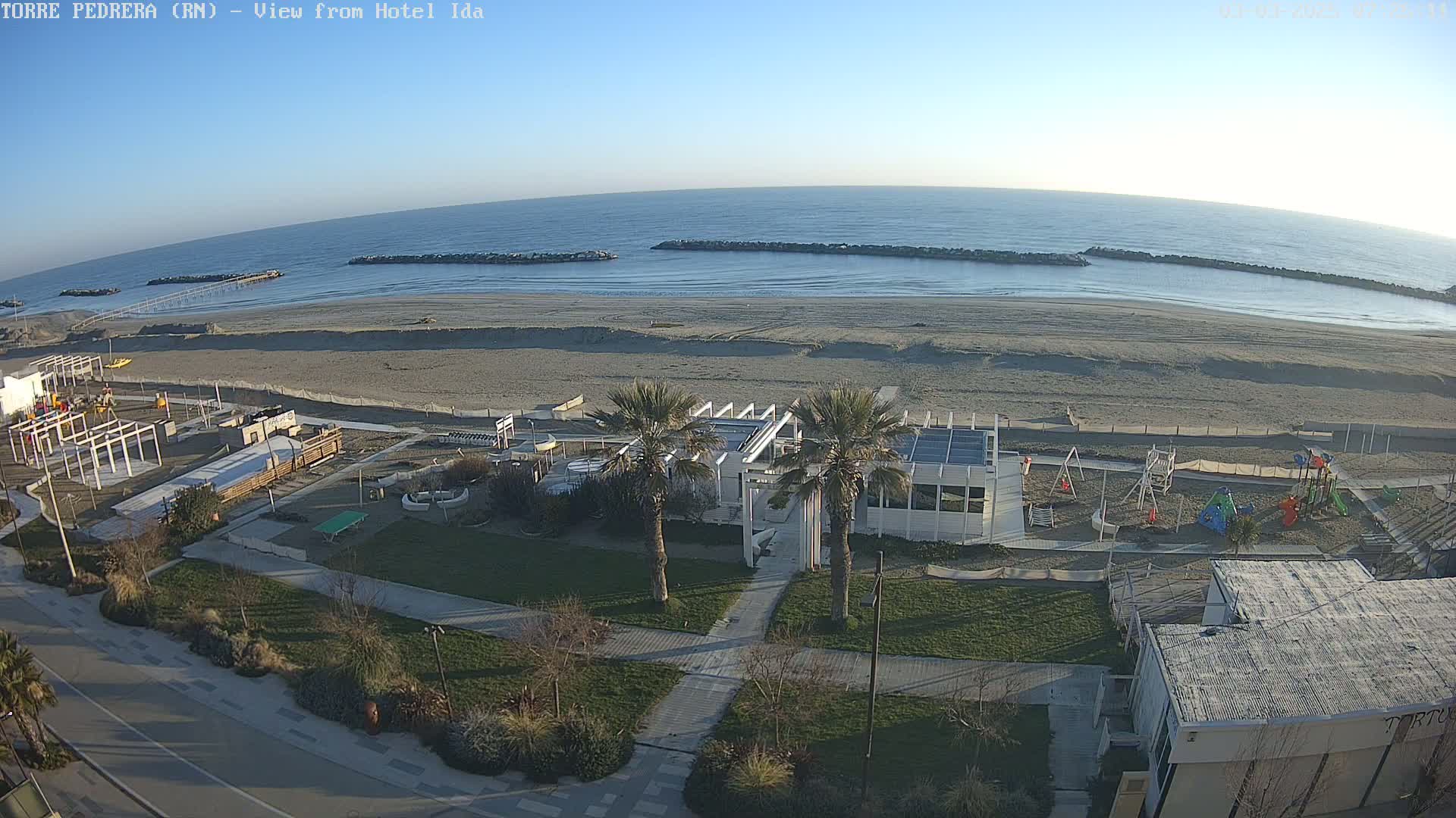 A beach with a calm ocean under a clear sky, featuring a playground, a beachfront building, and palm trees.
