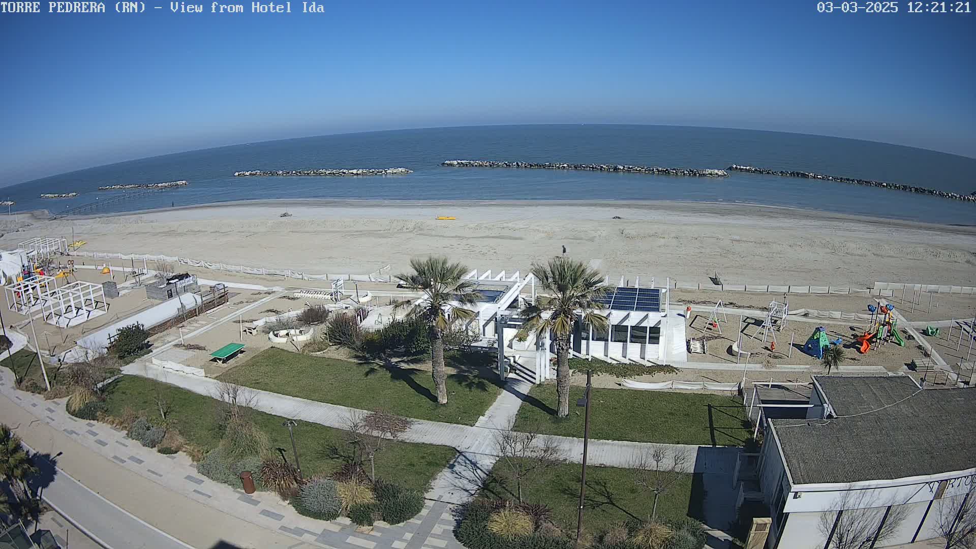 An aerial view of a sandy beach on a sunny day shows a playground, a building with solar panels, palm trees, and a walkway next to the beach, which is protected by breakwaters.