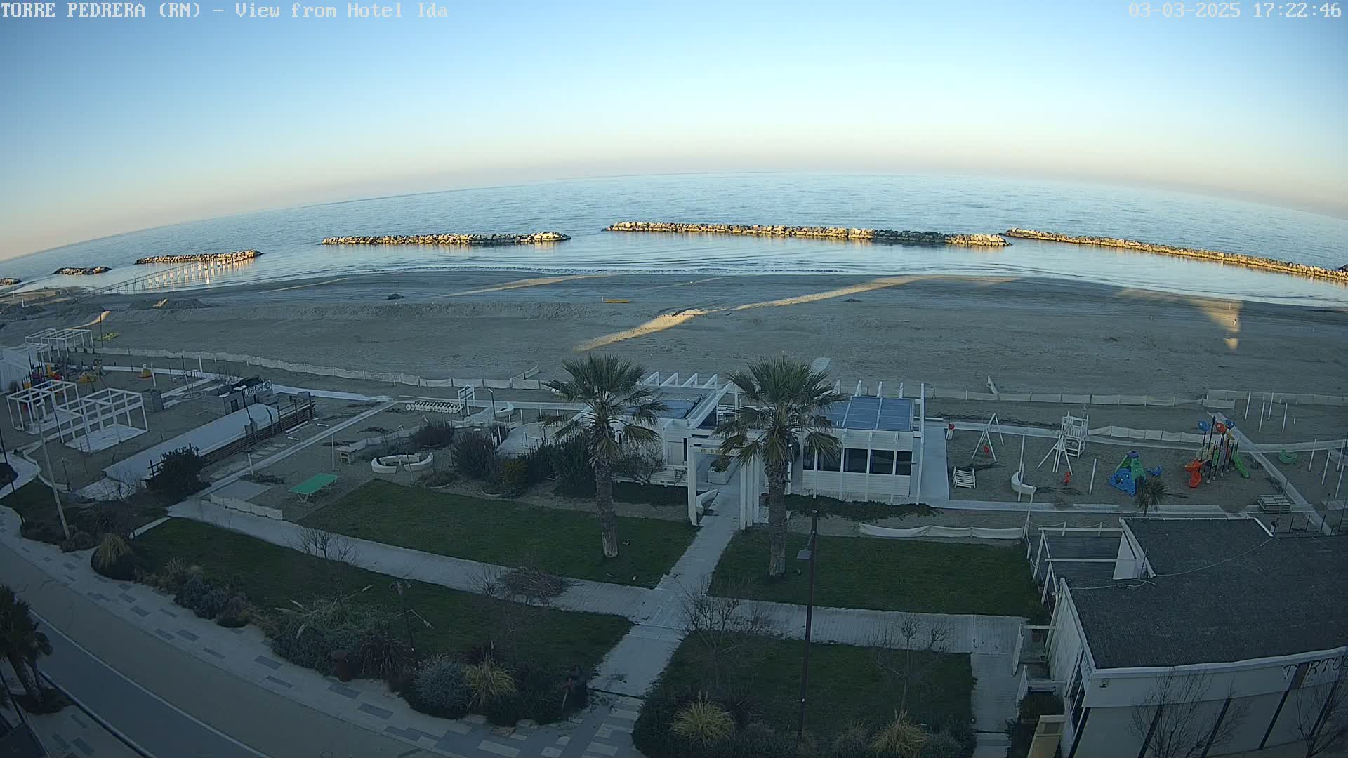 An aerial view shows a beach with calm water under a clear, twilight sky, featuring a beachfront building, playground, and palm trees.