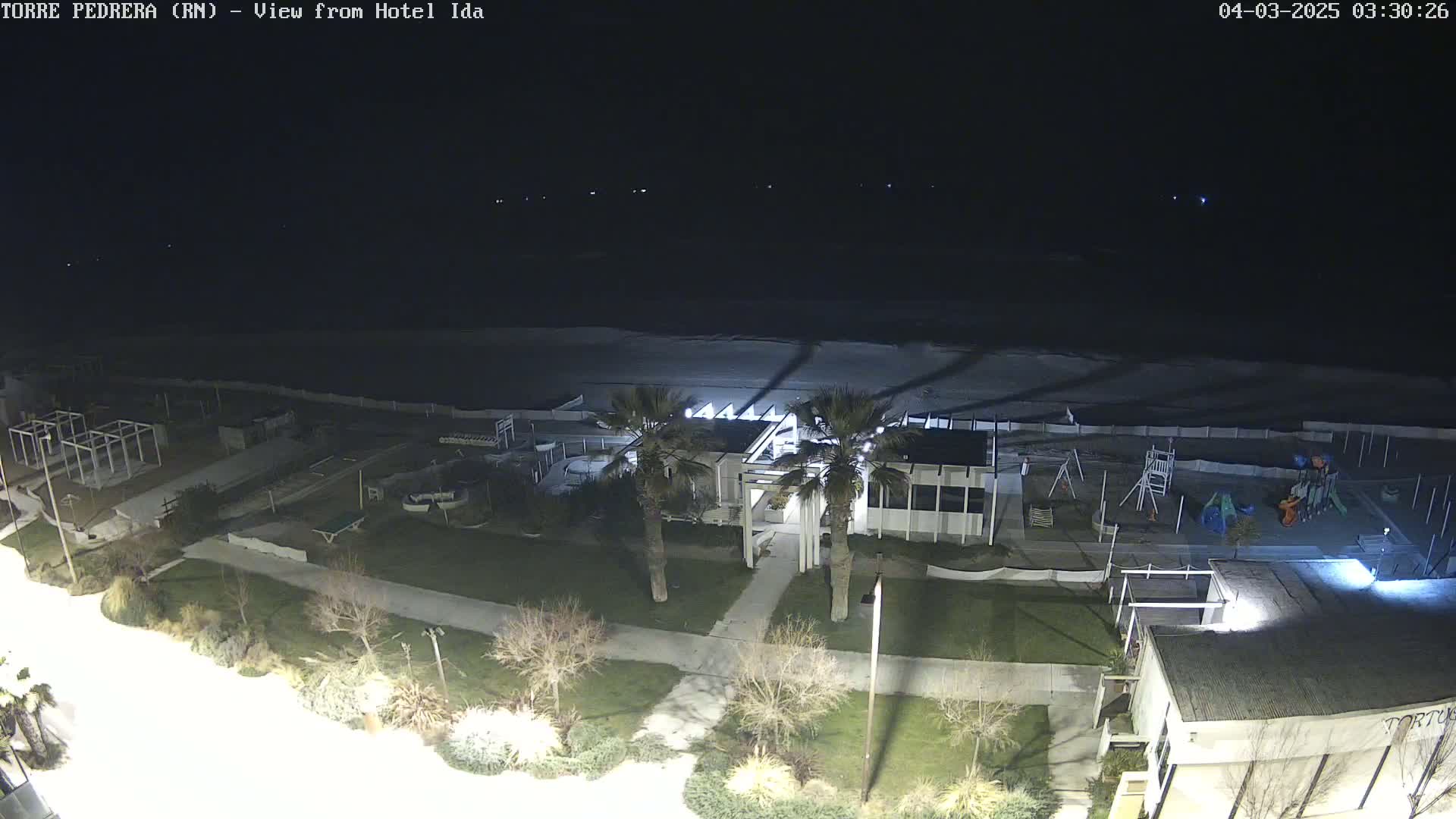 A nighttime aerial view of a beach area shows a building, playground, and a dark ocean under a clear sky.