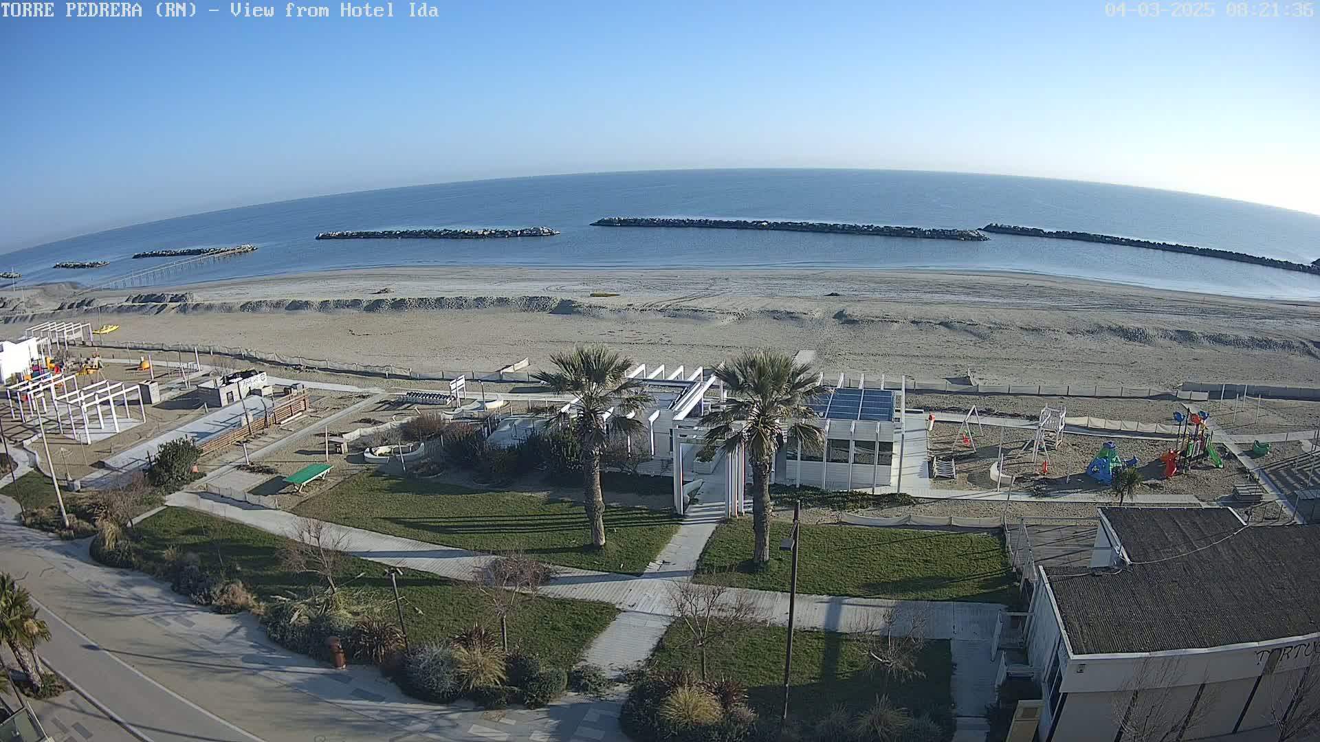A beach with calm, clear water under a sunny sky, featuring a playground, a building with solar panels, and palm trees on a grassy area next to a road.