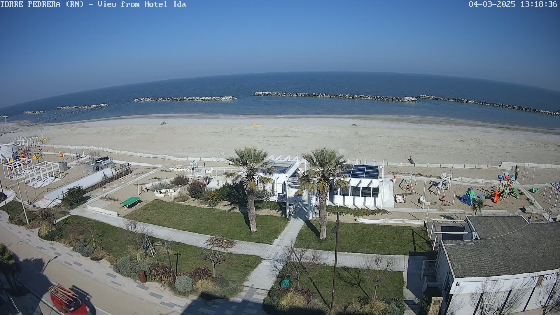 An aerial view shows a beach with a playground, a building with palm trees in front of it, and a calm sea under a clear blue sky.