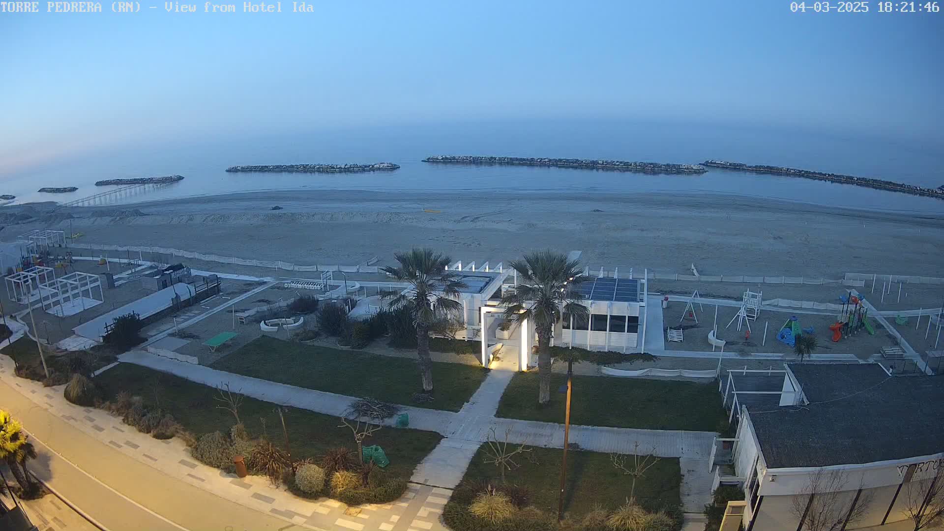 A beach with a playground, a beachfront building, and palm trees is visible at dusk under a clear, twilight sky.