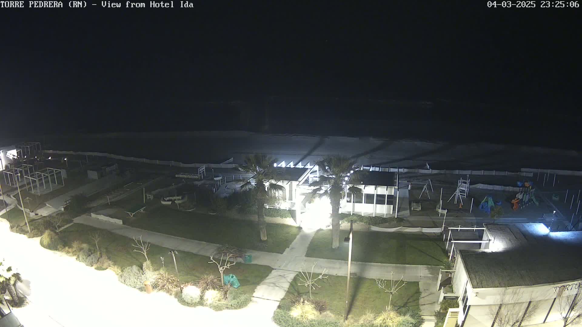 A nighttime aerial view of a beachside area shows a building, a playground, and a dark ocean under a clear sky.
