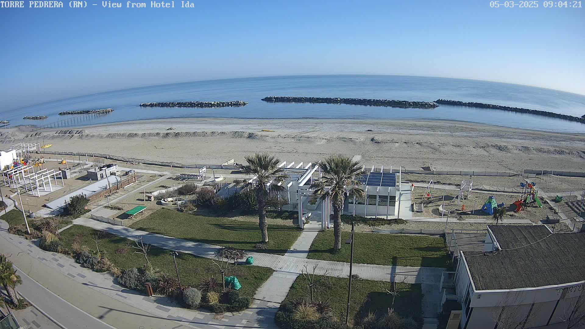 A beach with calm, blue water under a clear, sunny sky is viewed from above, showing a playground, grassy areas, and palm trees near a building.