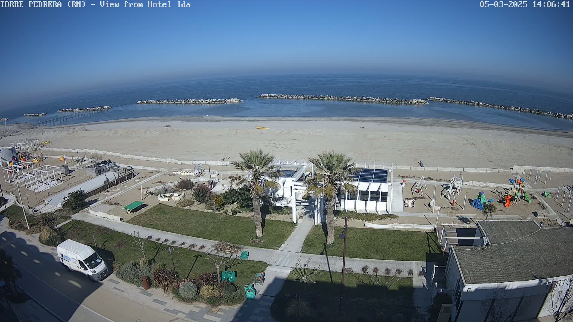 A beachfront view on a sunny day shows a sandy beach with calm water, a playground, a building with solar panels, and palm trees lining a paved walkway.