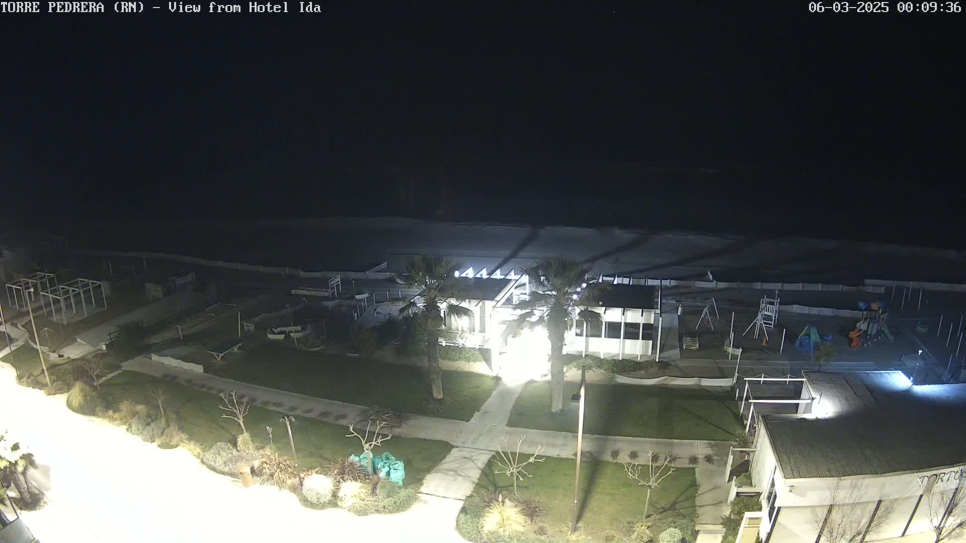 A nighttime view of a beach, playground, and building complex, illuminated by artificial lights, with calm waves visible in the distance.
