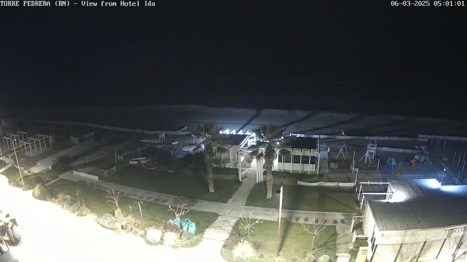 A nighttime, high-angle view shows a beach, a playground, and a building with palm trees, all illuminated under a dark sky.