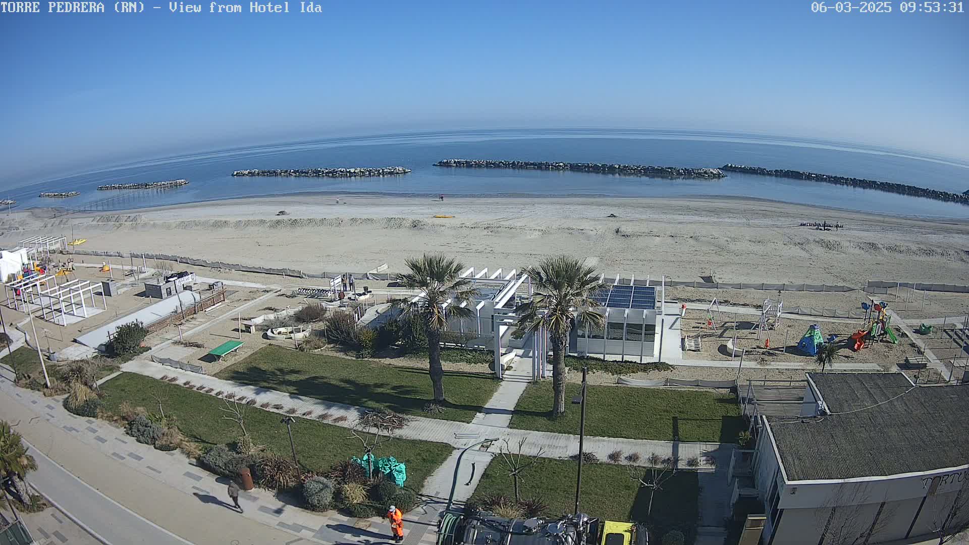 A sandy beach with calm, clear water under a sunny sky is seen from above, along with adjacent grassy areas containing playgrounds, palm trees, and buildings.
