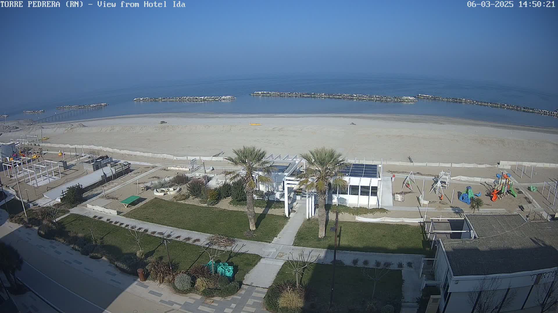 A beach with a playground and a small building is visible, next to a calm sea under a clear blue sky.