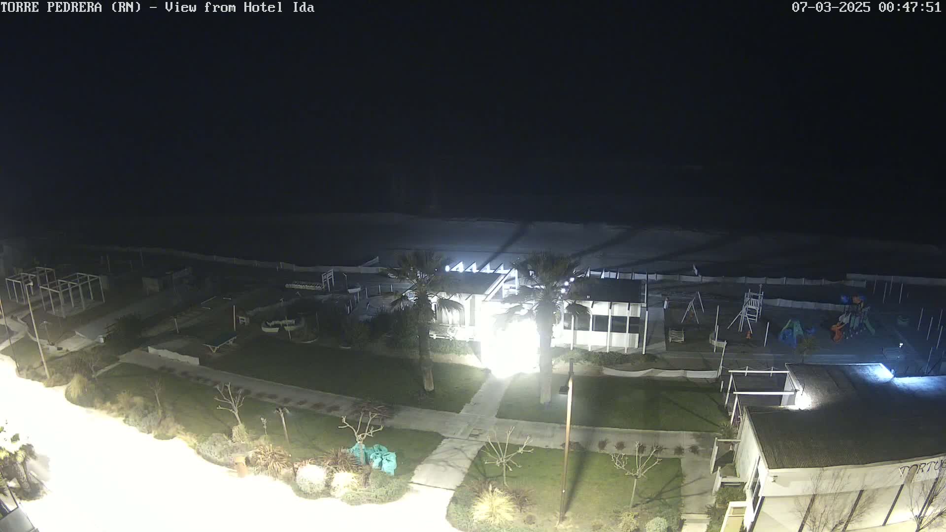 A nighttime view of a beach, a playground, and a building, with palm trees and landscaping visible, under a dark sky.