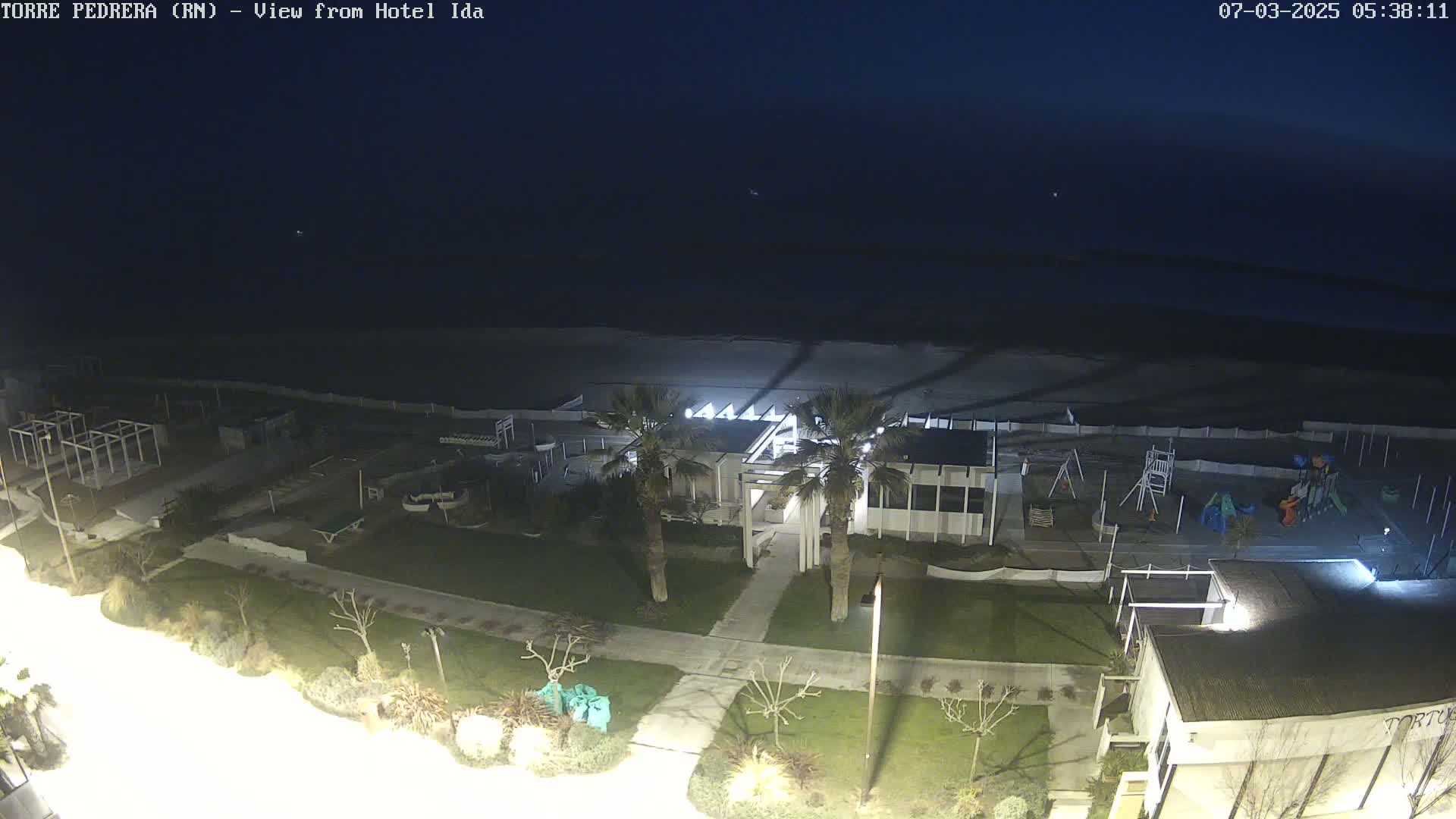 A nighttime aerial view shows a beach, a playground, and a building complex illuminated by artificial lights, under a dark, clear sky.
