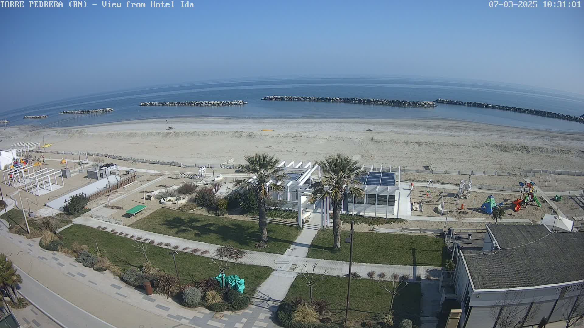 A beach with a playground and a white building is shown on a sunny day, with calm, blue water visible in the background.