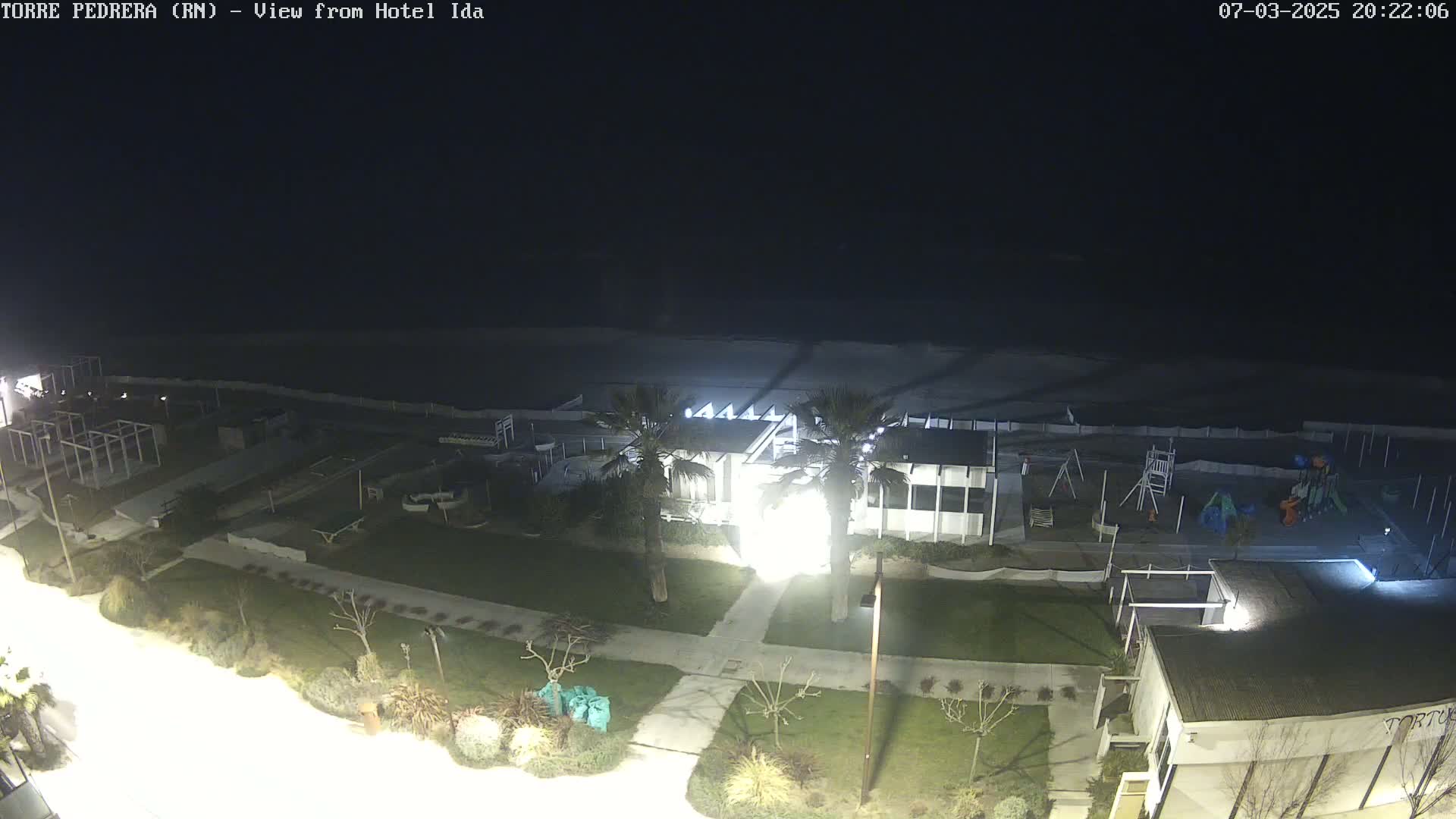 A nighttime aerial view of a beachfront area shows a building, playground, and beach with calm water under a dark sky.