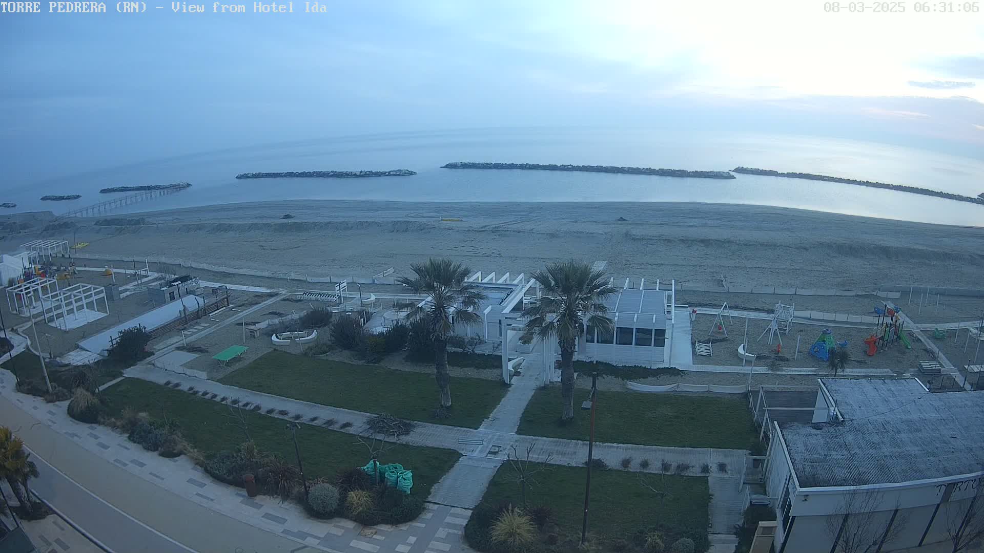 An overcast day shows a beach with a playground, beachside buildings, and palm trees, all viewed from above.