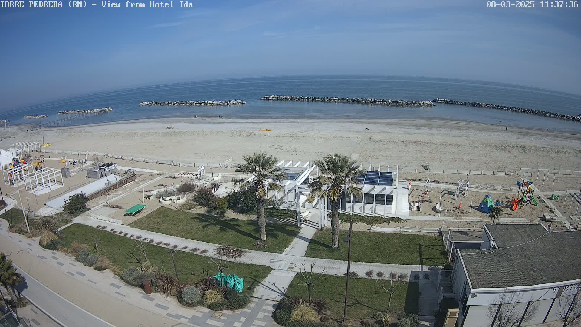 An aerial view shows a sandy beach on a sunny day, with a playground, palm trees, and a building near the shoreline.